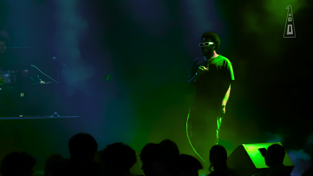 Person standing on stage with microphone, audience silhouettes in foreground, green and blue lighting, dark background.