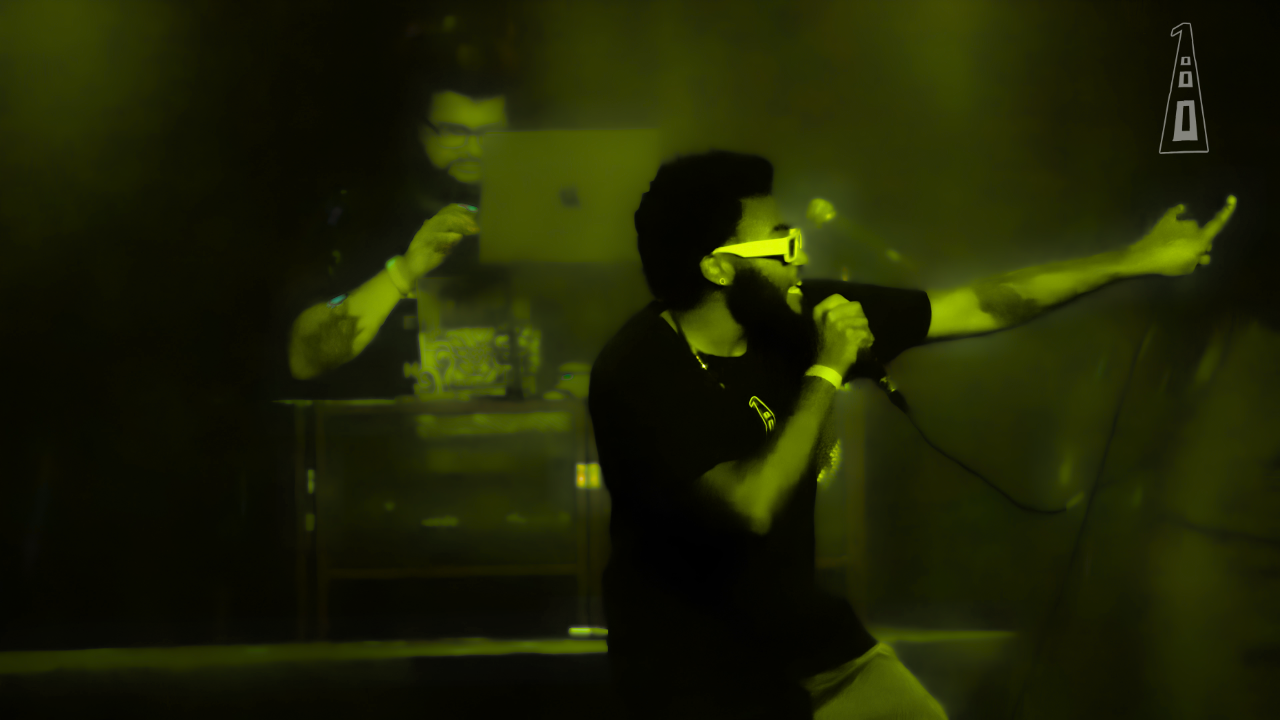 Person singing into a microphone on stage with a person in the background using a laptop, both in a dark setting.