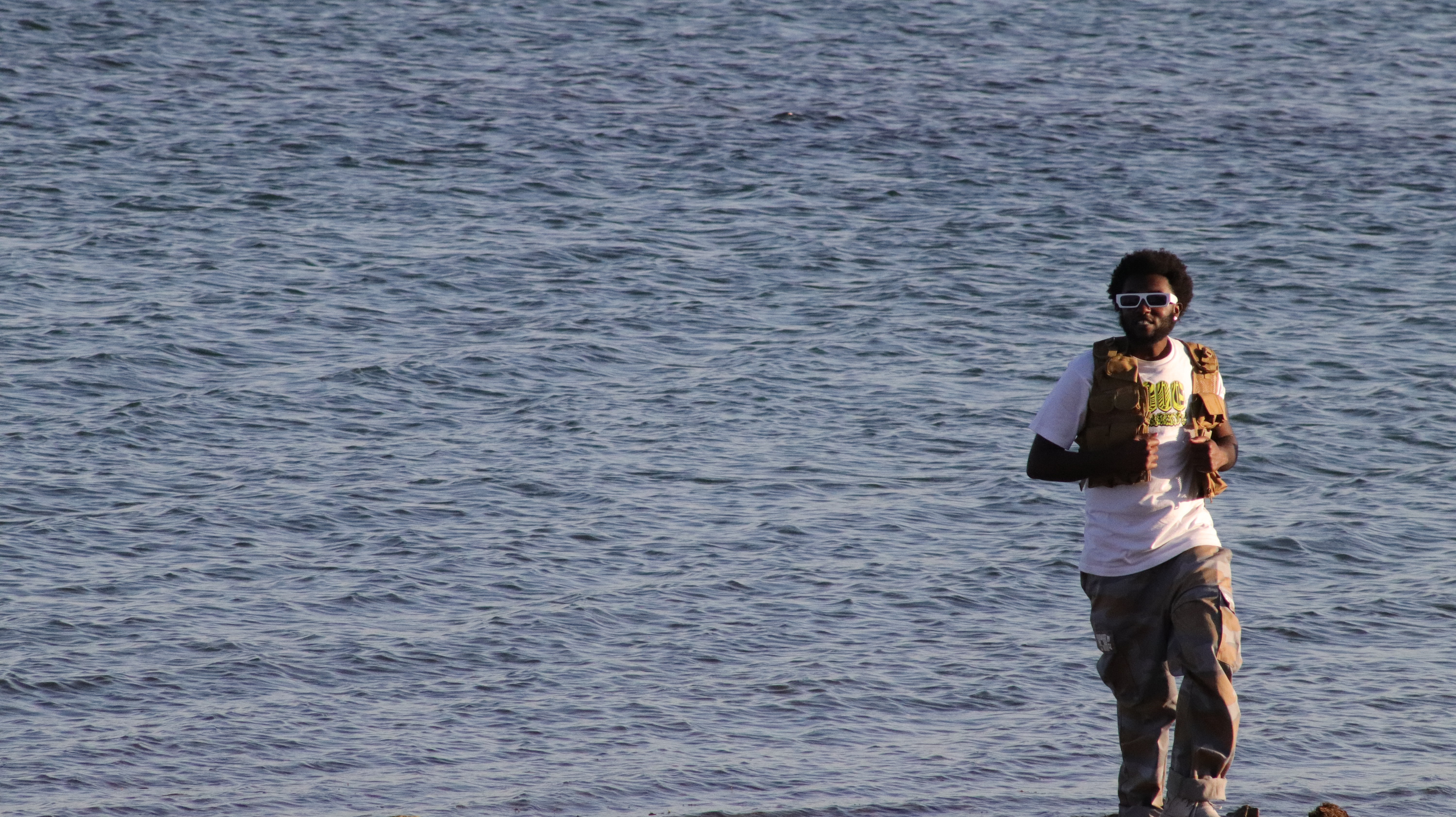 Person walking along water's edge holding a yellow object, wearing a white shirt and dark pants.