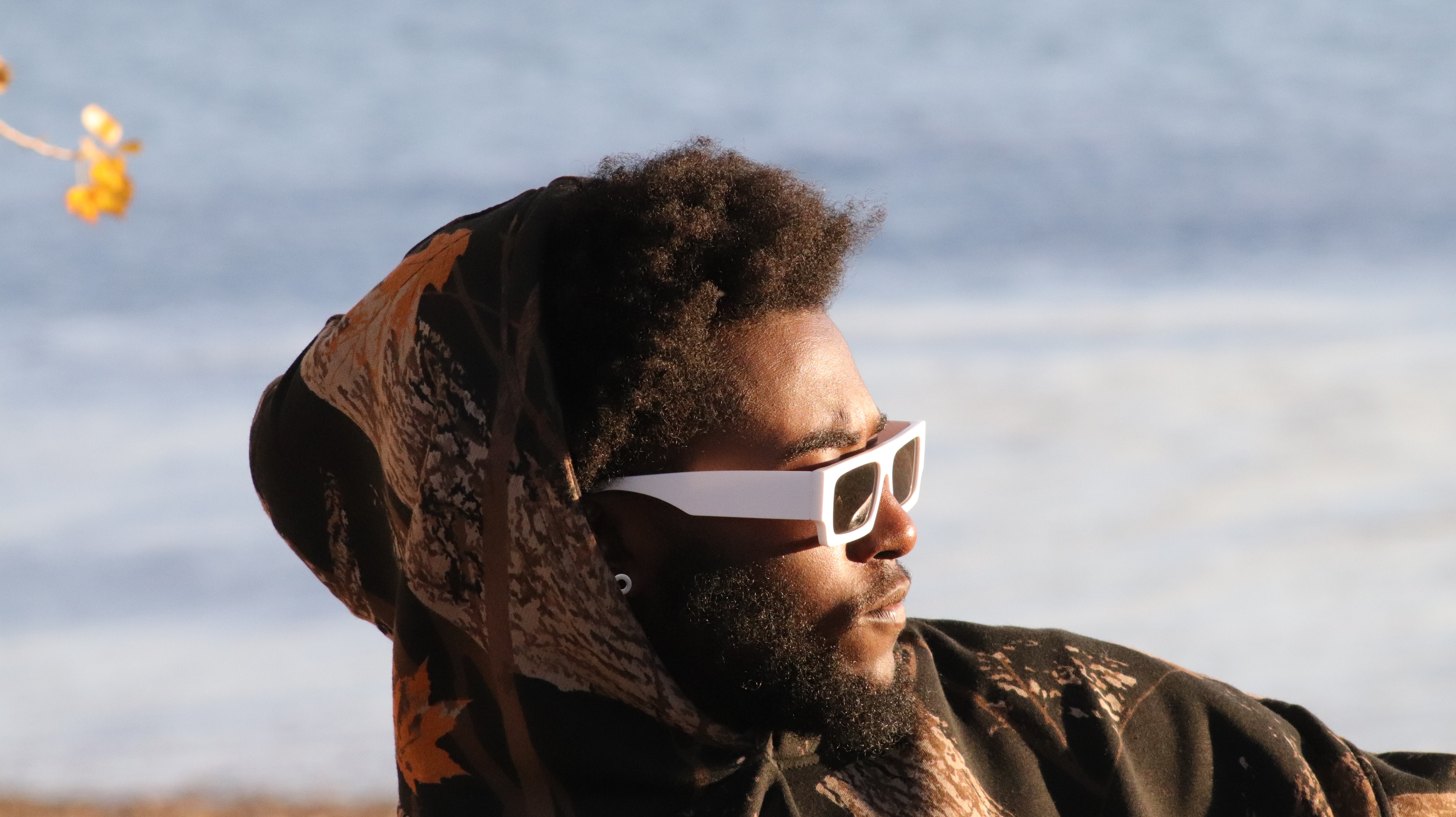 Person with curly hair wearing sunglasses and a hood, looking to the side near a body of water.