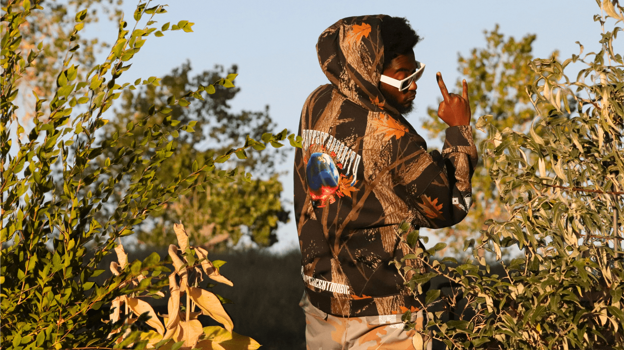 Person wearing a hoodie and sunglasses standing among trees and bushes outdoors.