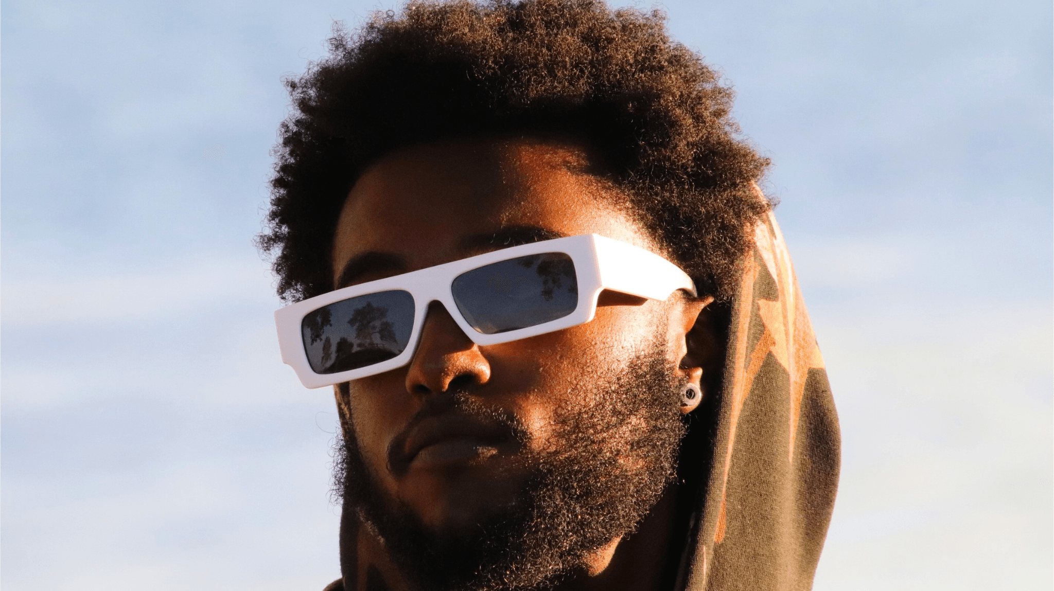 Person with curly hair wearing white sunglasses outdoors, sky in background.