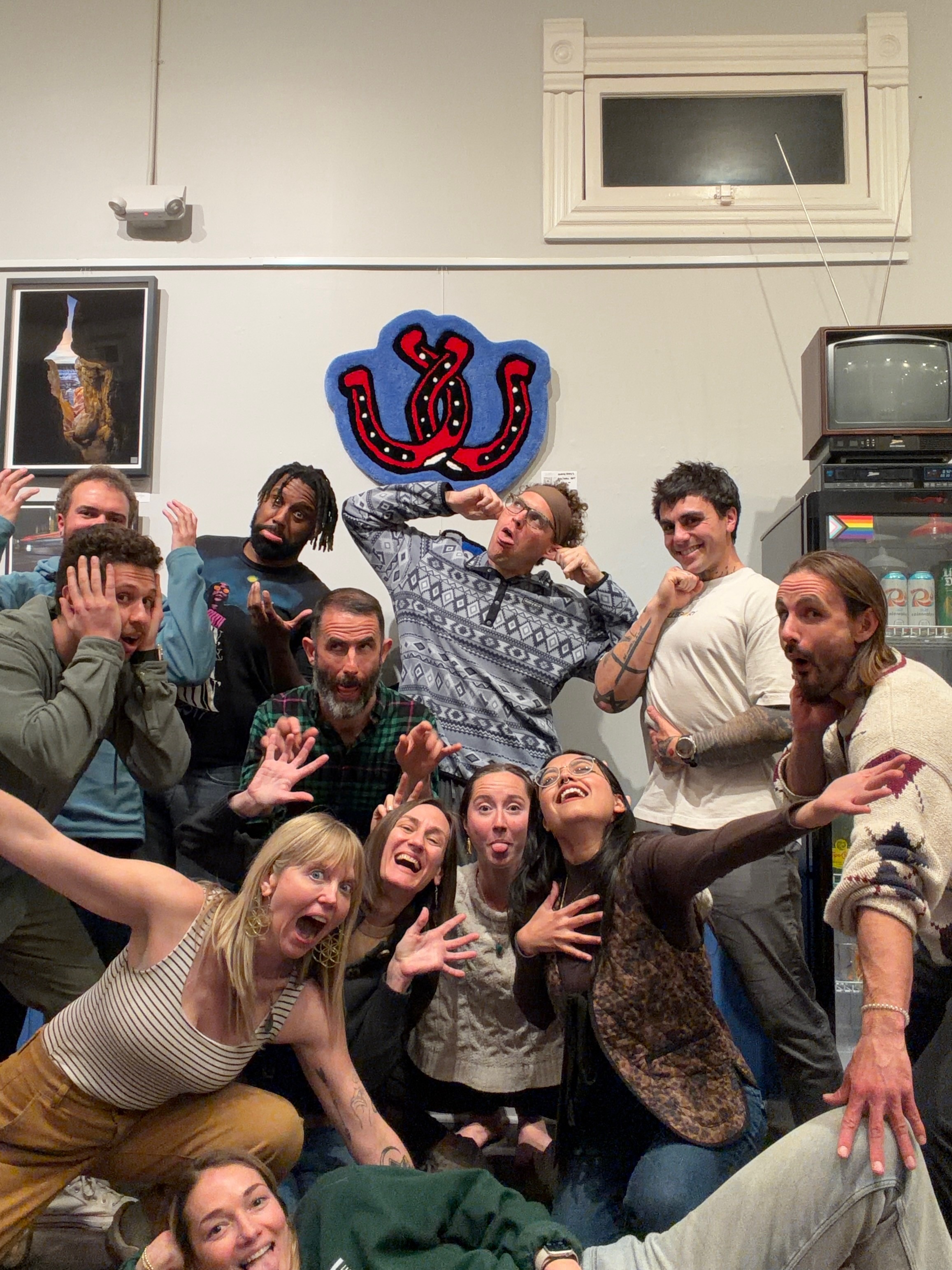 Group of people posing indoors with a colorful wall decoration, some making gestures, smiling, and looking at the camera.