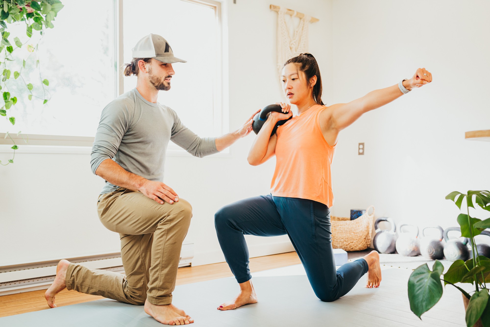 Woman practicing boxing with trainer in bright room, woman in orange top and blue pants, trainer in gray shirt and cap, woman in a lunge position