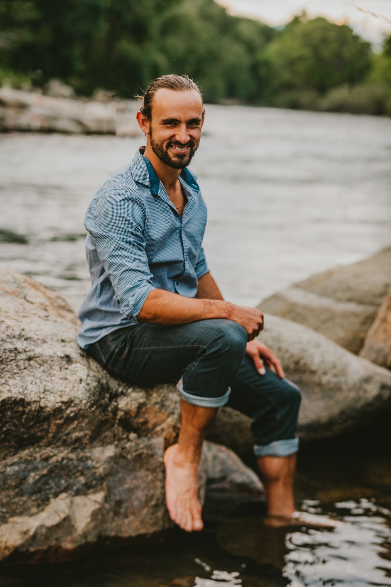 Man sitting on a rock by a river, smiling, with trees and water in the background.