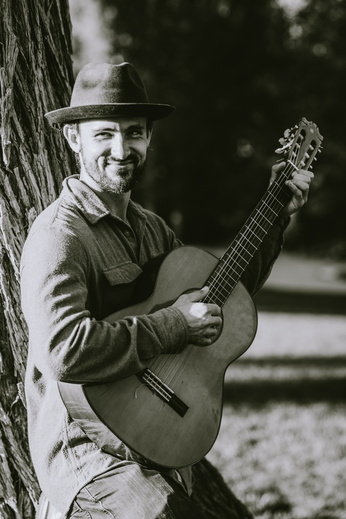 Man with a hat holding an acoustic guitar outdoors, smiling, standing next to a tree, in black and white.