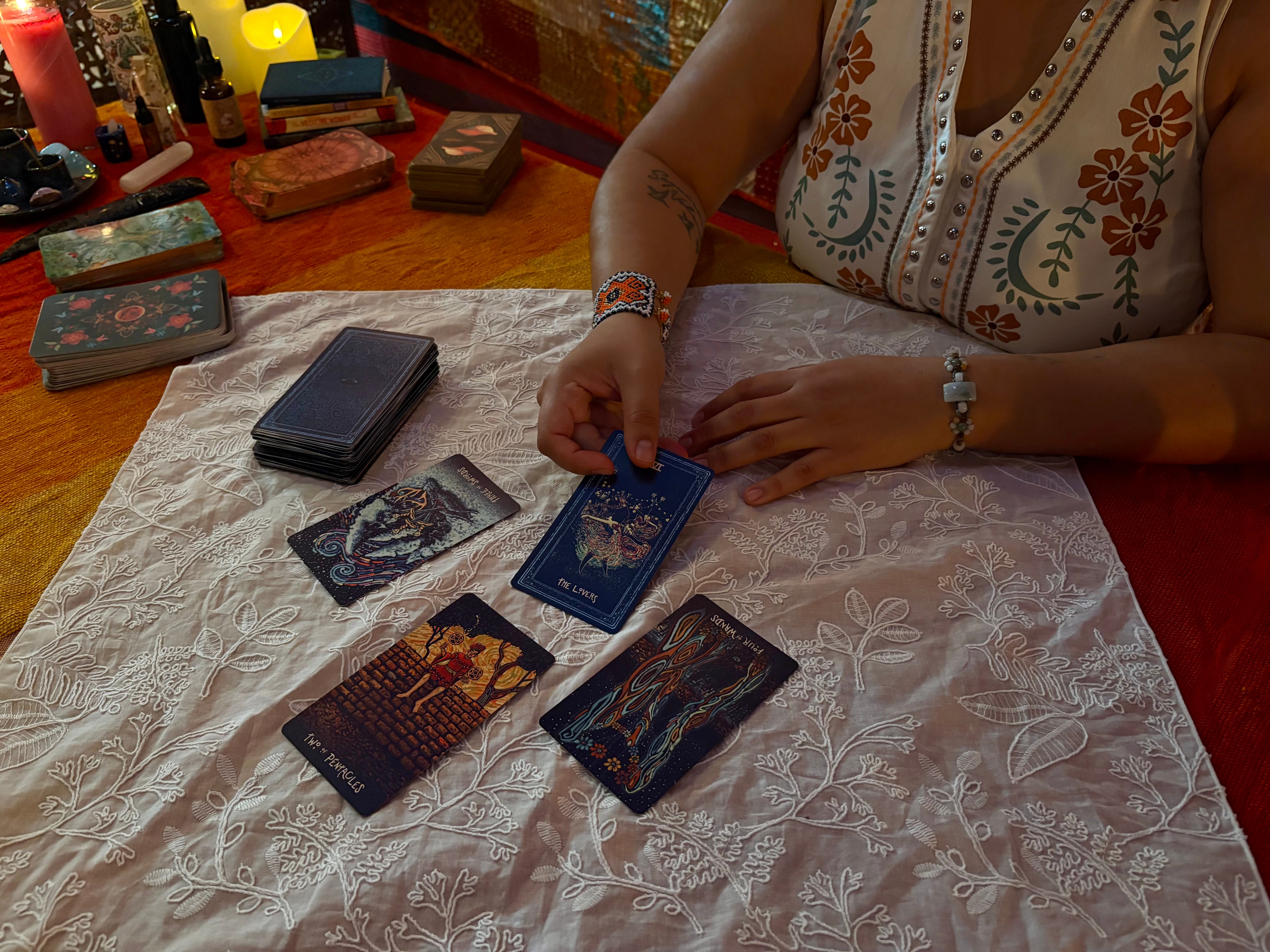 Person in embroidered top holding tarot cards on a table with other cards and boxes, lace tablecloth, and colorful background.
