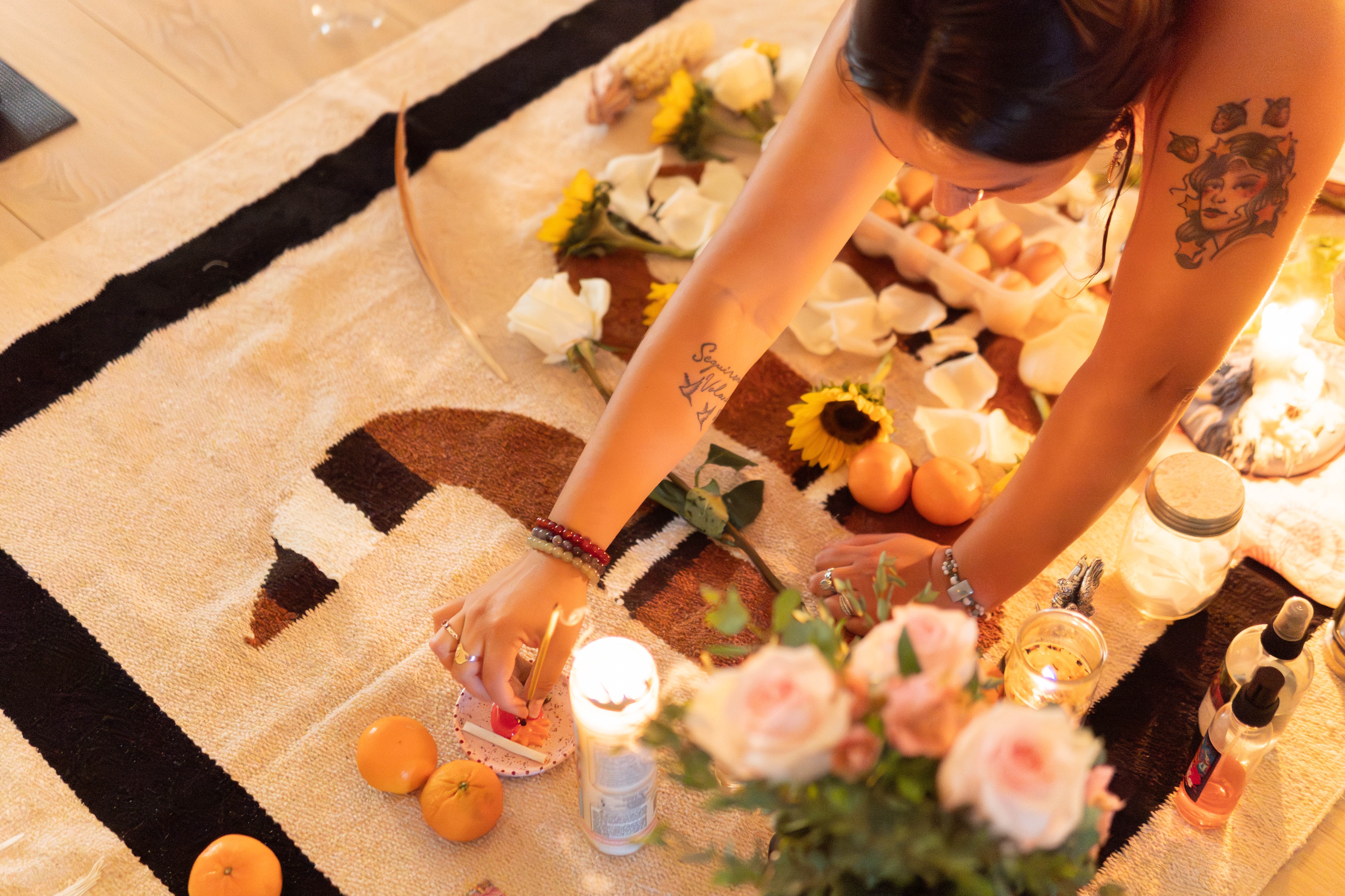 Person with tattoos arranging flowers and candles on a patterned rug, surrounded by flowers and decorative items.