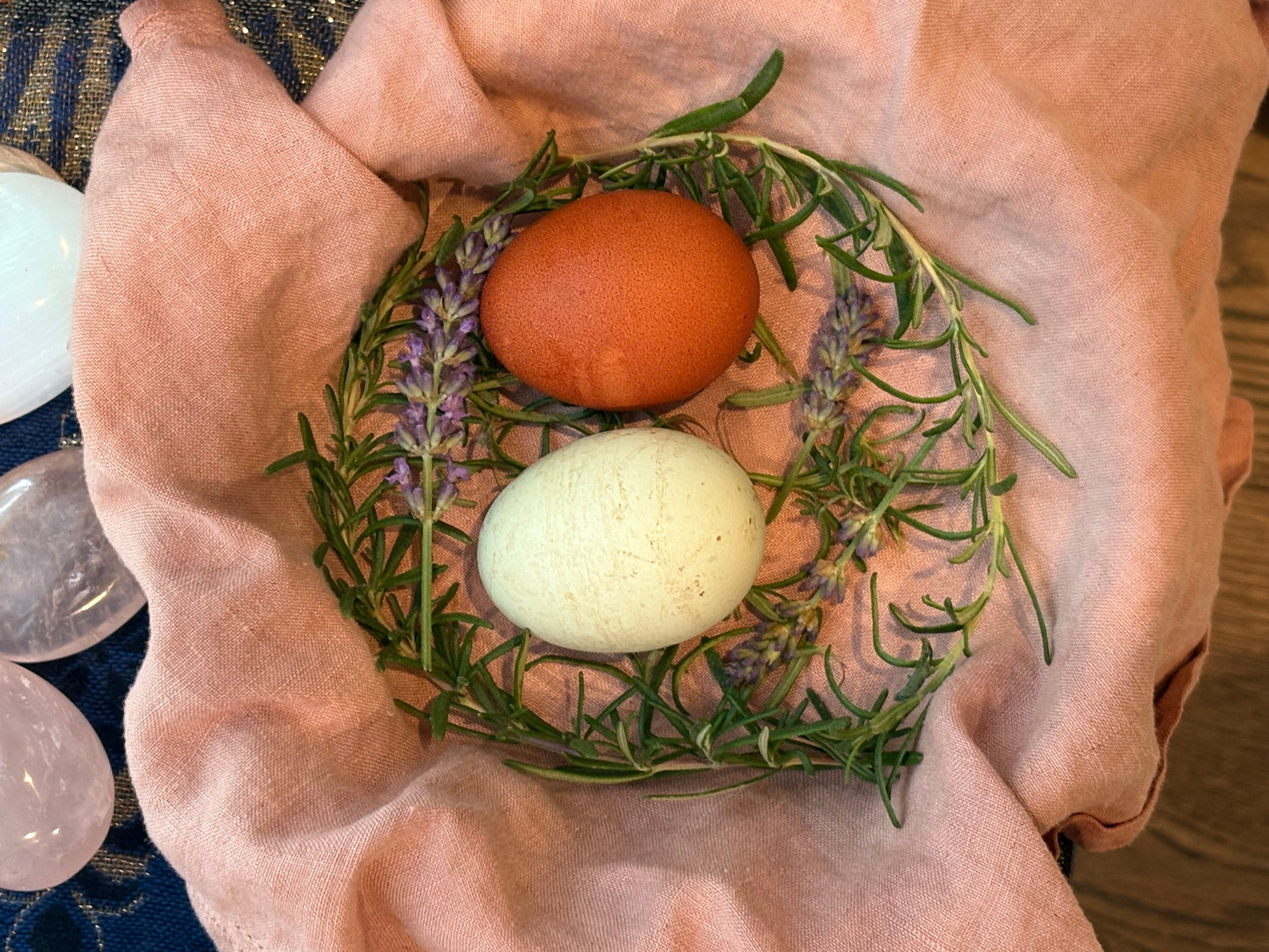 Two eggs, one brown and one white, are surrounded by green and purple herbs on a pink cloth.