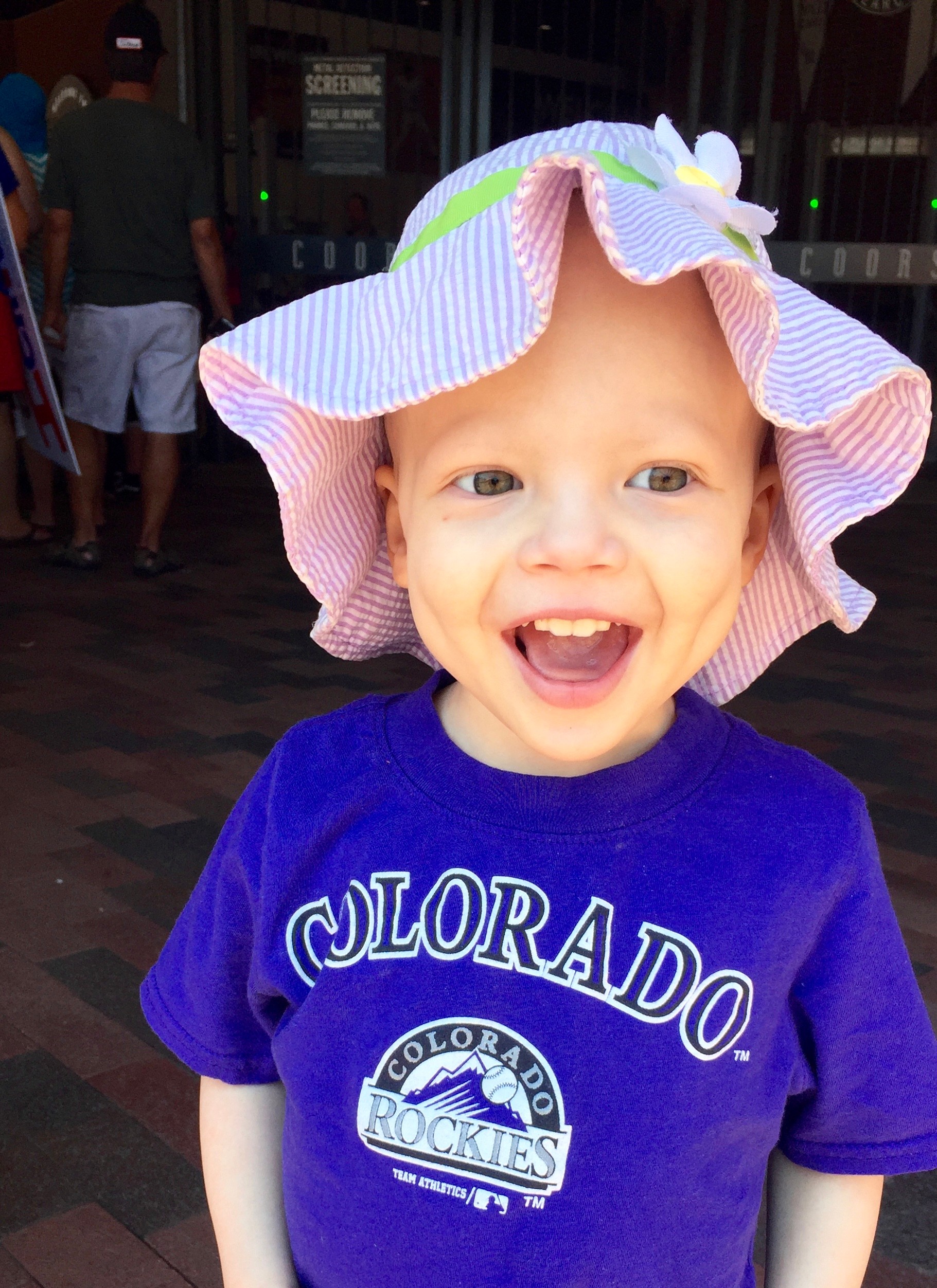 Smiling young child wearing a purple Colorado Rockies shirt and a colorful hat, indoors with people in the background.