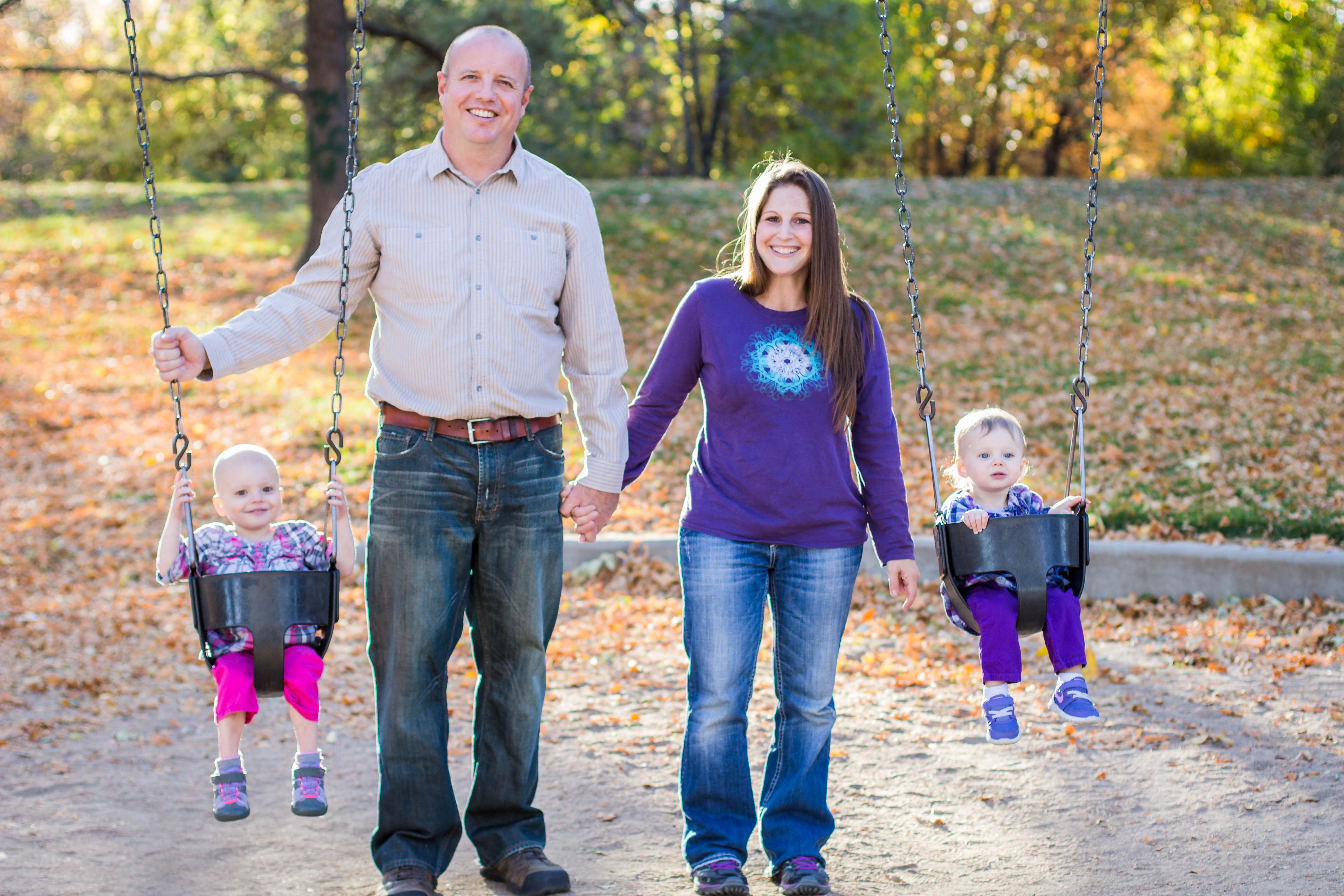 A family of four at a park, two children on swings, holding hands, autumn trees in background.