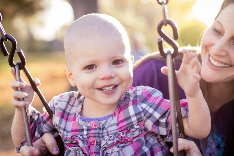 Smiling baby on a swing with a woman in the background outdoors, sunny day, blurred trees, and warm lighting.