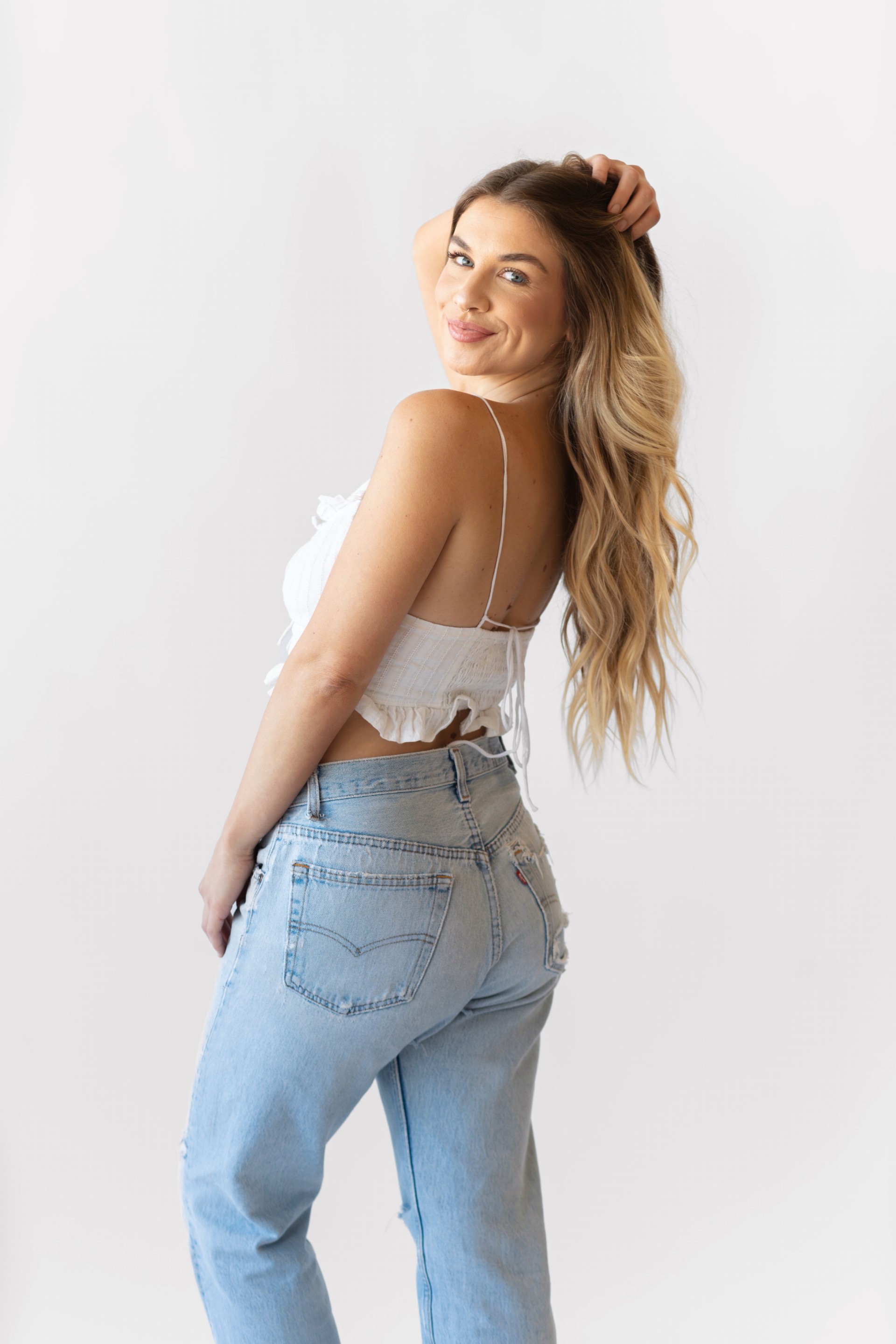 Young woman with long wavy hair smiling, wearing a white top and light blue jeans, standing against a plain white background.