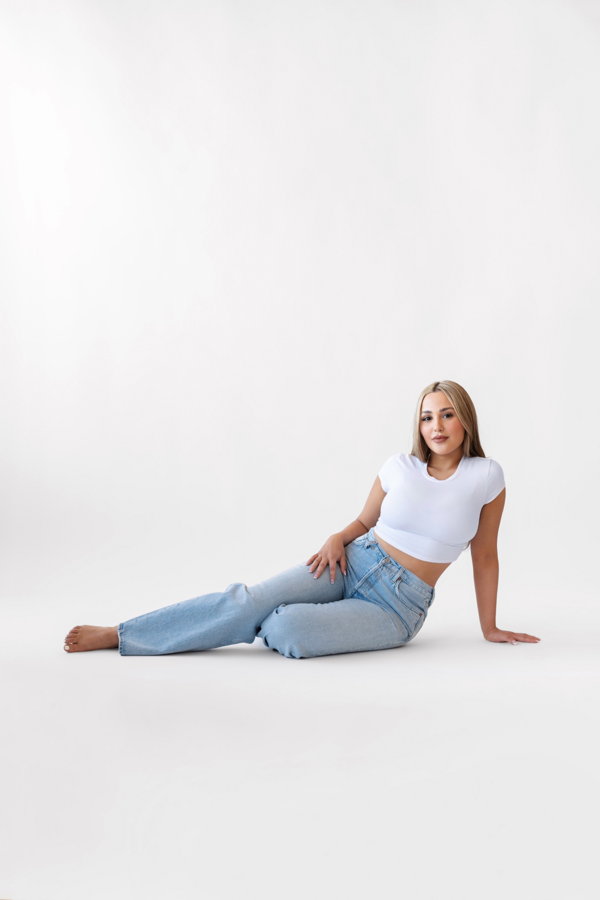 Young woman with blonde hair sitting on the floor, wearing a white crop top and light blue jeans, against a plain white background.