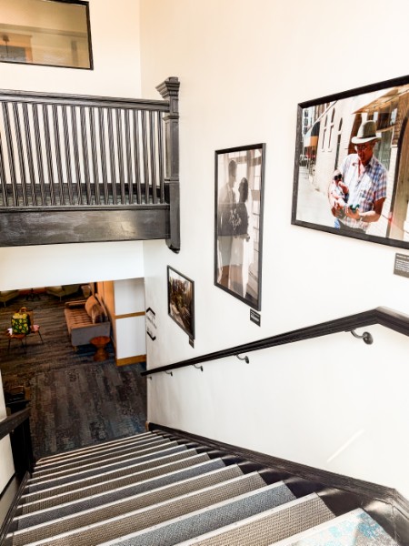 Staircase viewed from above with framed photographs on white wall, wooden railing, and a glimpse of a room below.