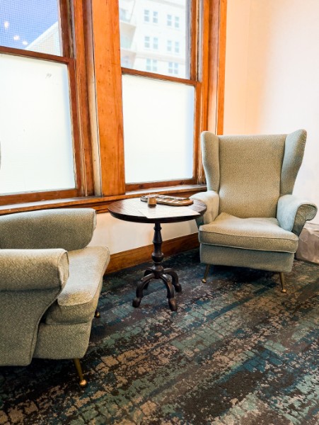 Two armchairs and a small round table near a window with wooden frames, sunlight coming in, patterned carpet on the floor.