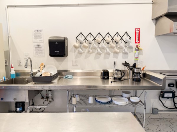 Clean kitchen counter with dishes, utensils, and kitchen tools, with wall-mounted hooks and a paper towel dispenser, in a commercial setting.