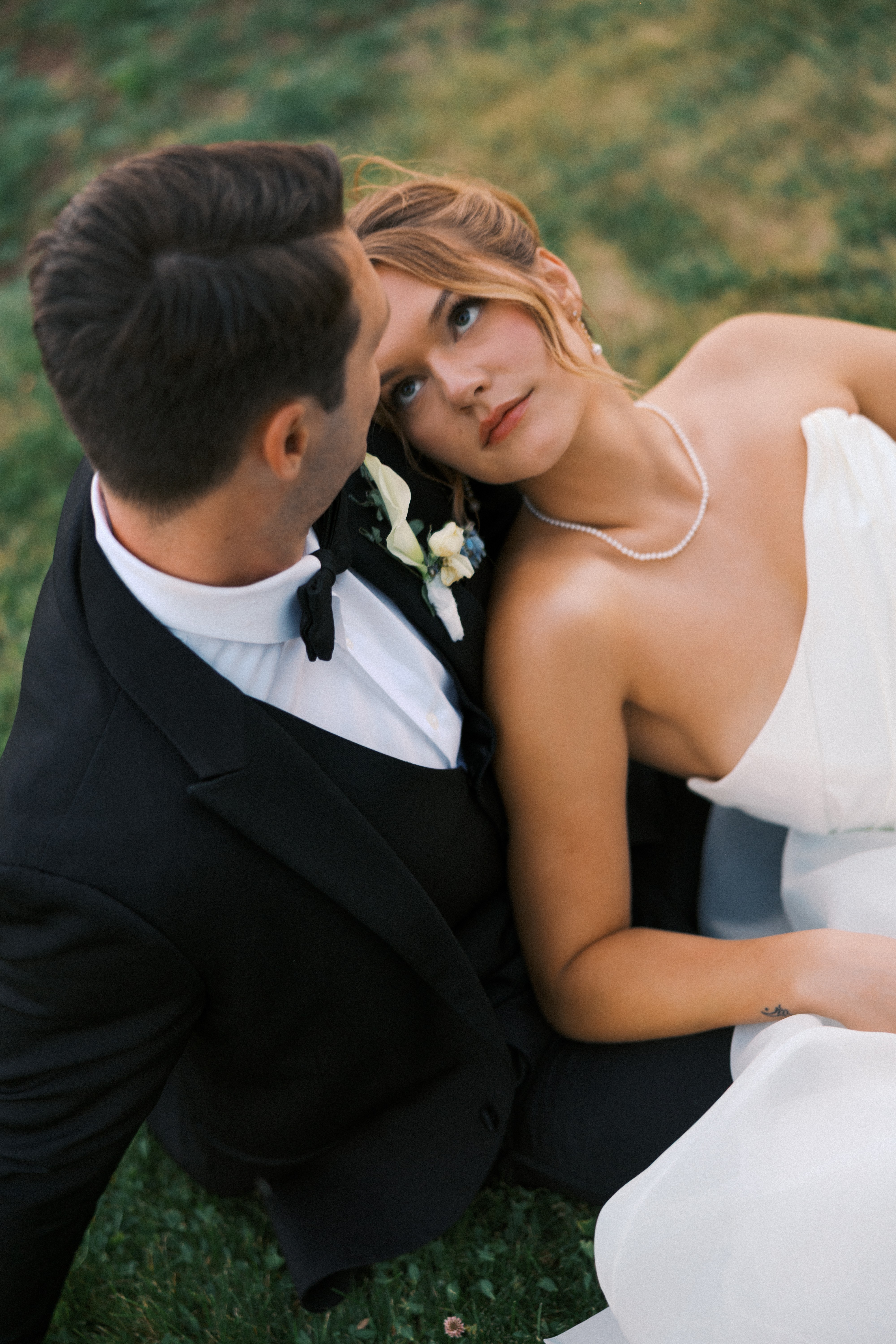 A couple in wedding attire sitting outdoors, gazing at each other, with a blurred green background.