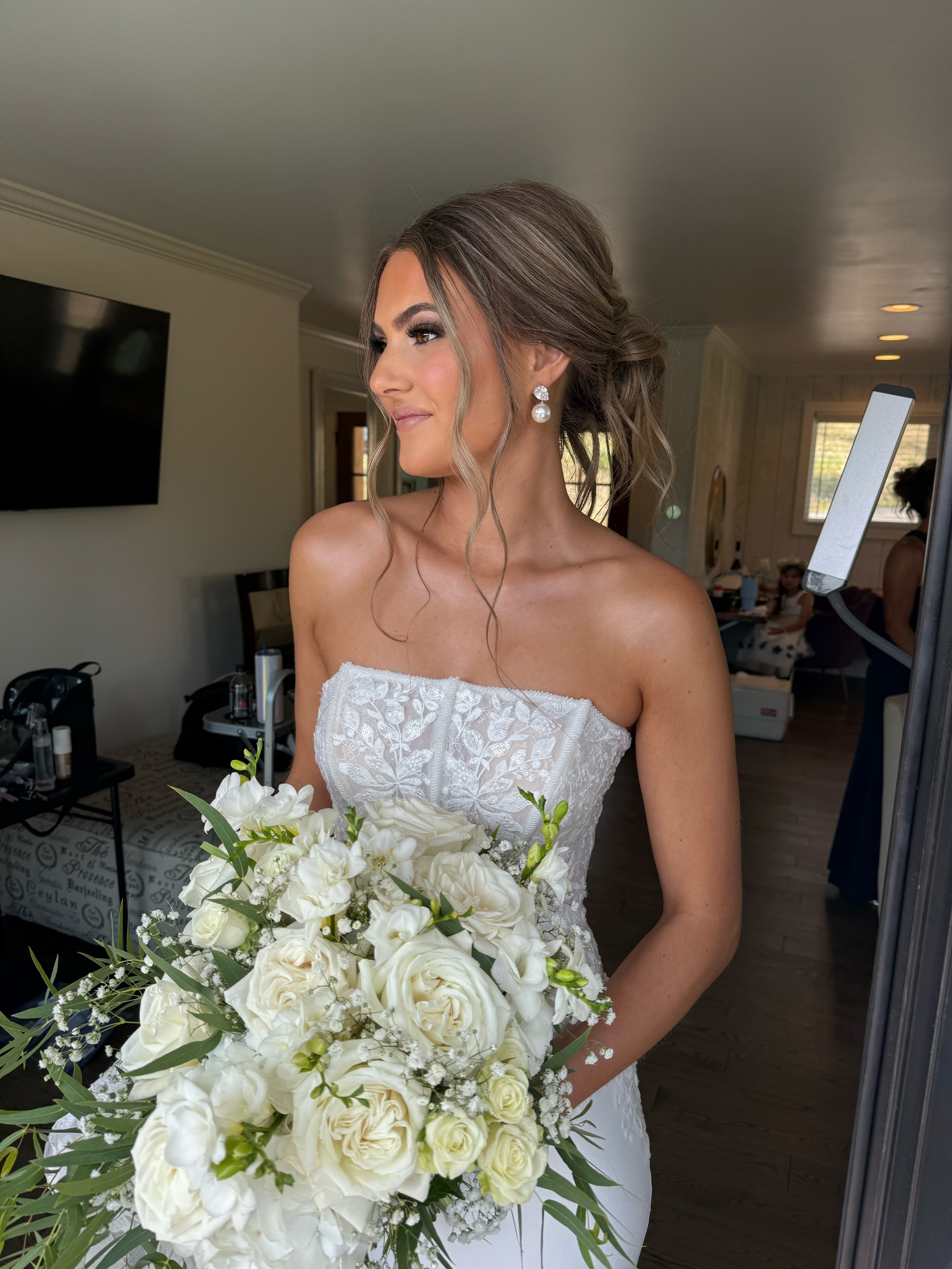 Woman in wedding dress holding a large bouquet of white flowers, smiling, indoors with a TV on the wall.