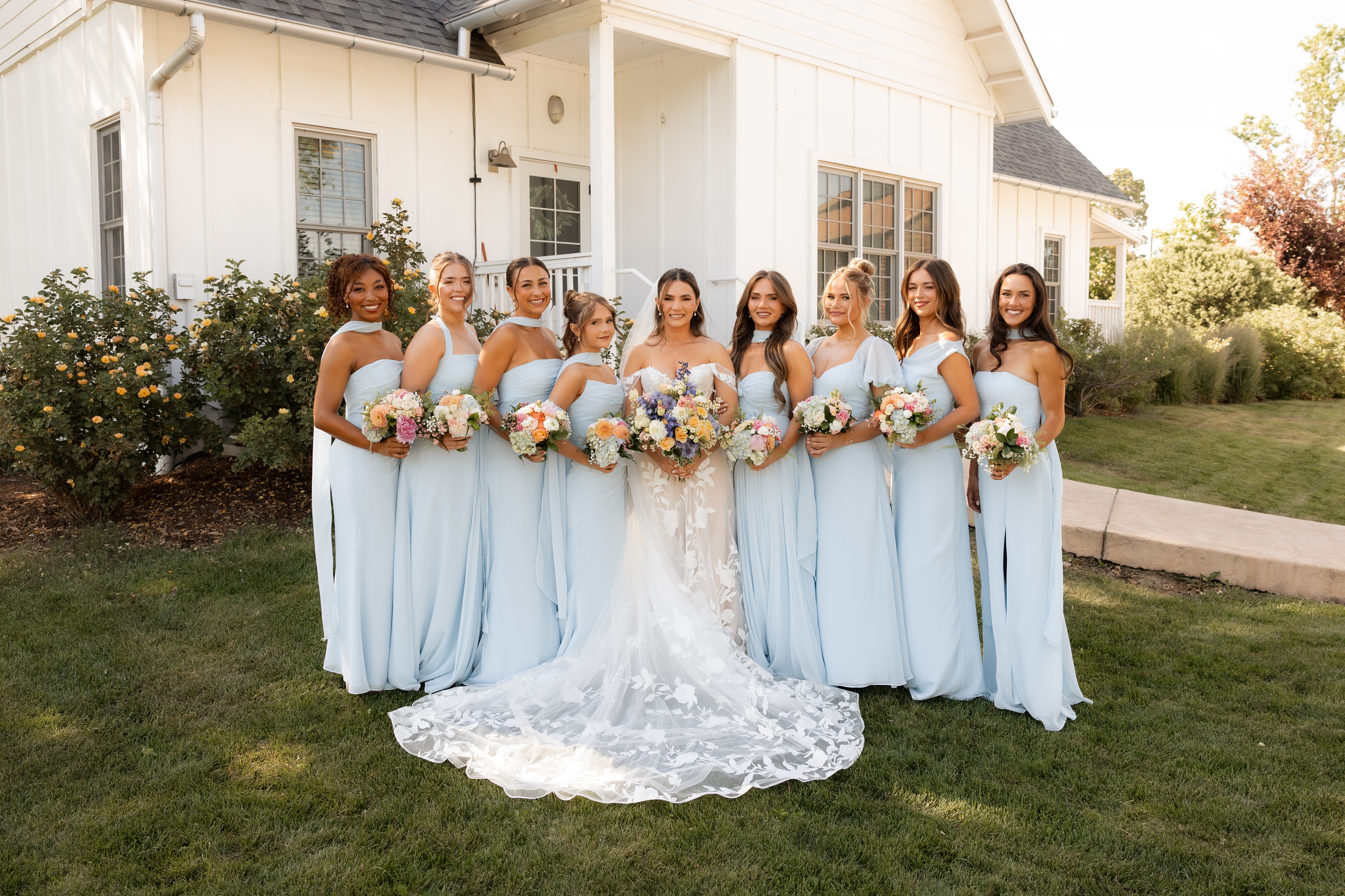 Group of eight women in light blue dresses, one in a white wedding gown, standing outdoors in front of a house, holding bouquets.