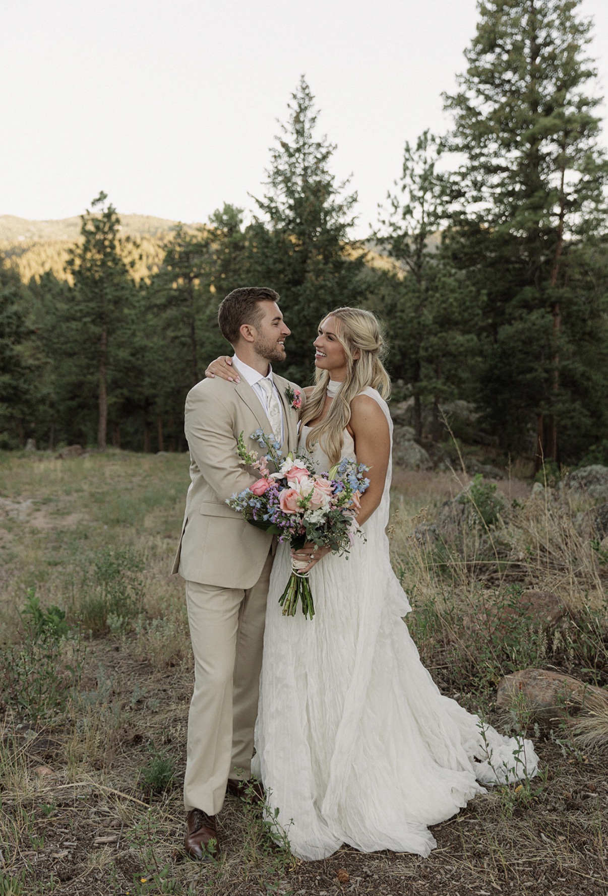 A bride and groom stand outdoors in a forested area, gazing at each other, with trees and mountains in the background.