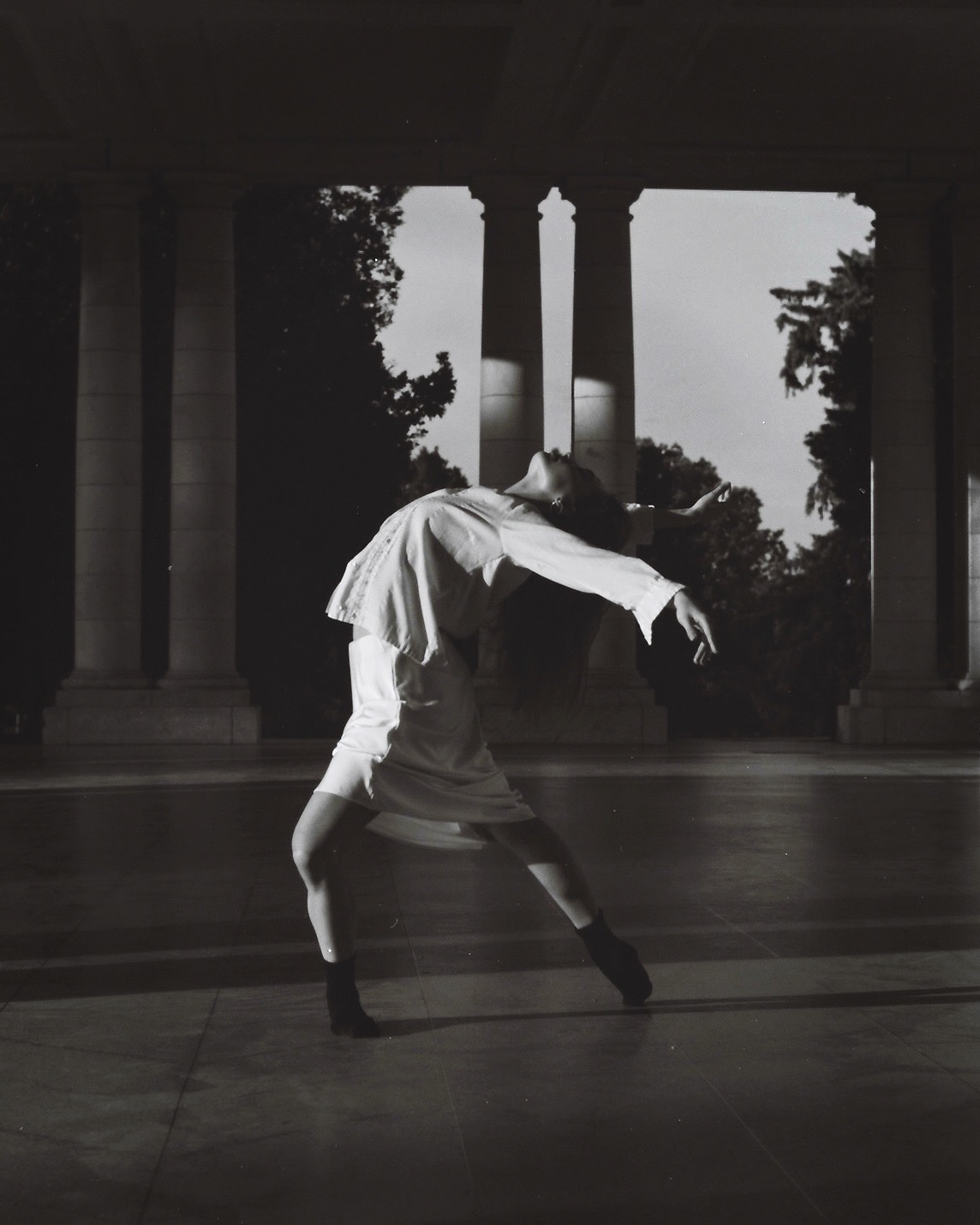 Person performing a backbend dance move indoors with columns and trees outside in the background.