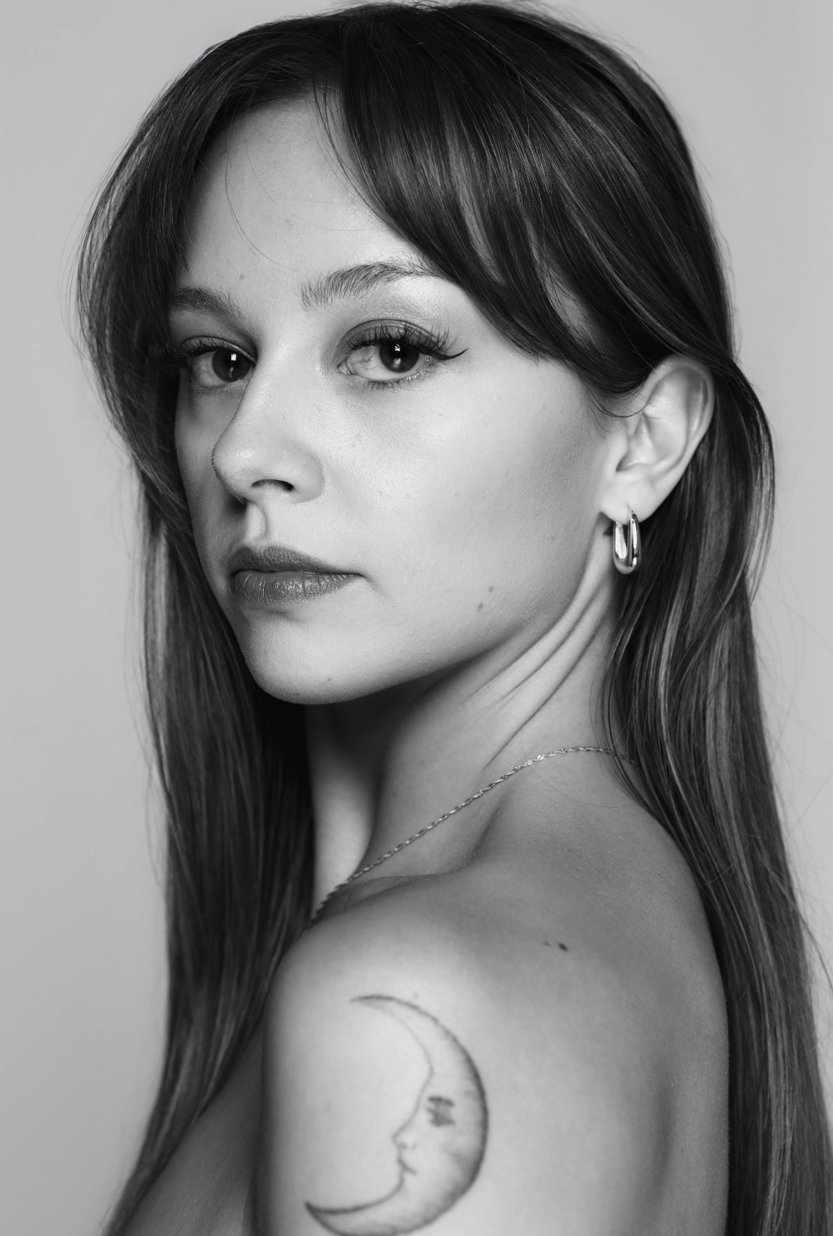Black and white portrait of a woman with long hair, looking sideways, showing her shoulder and a moon tattoo.
