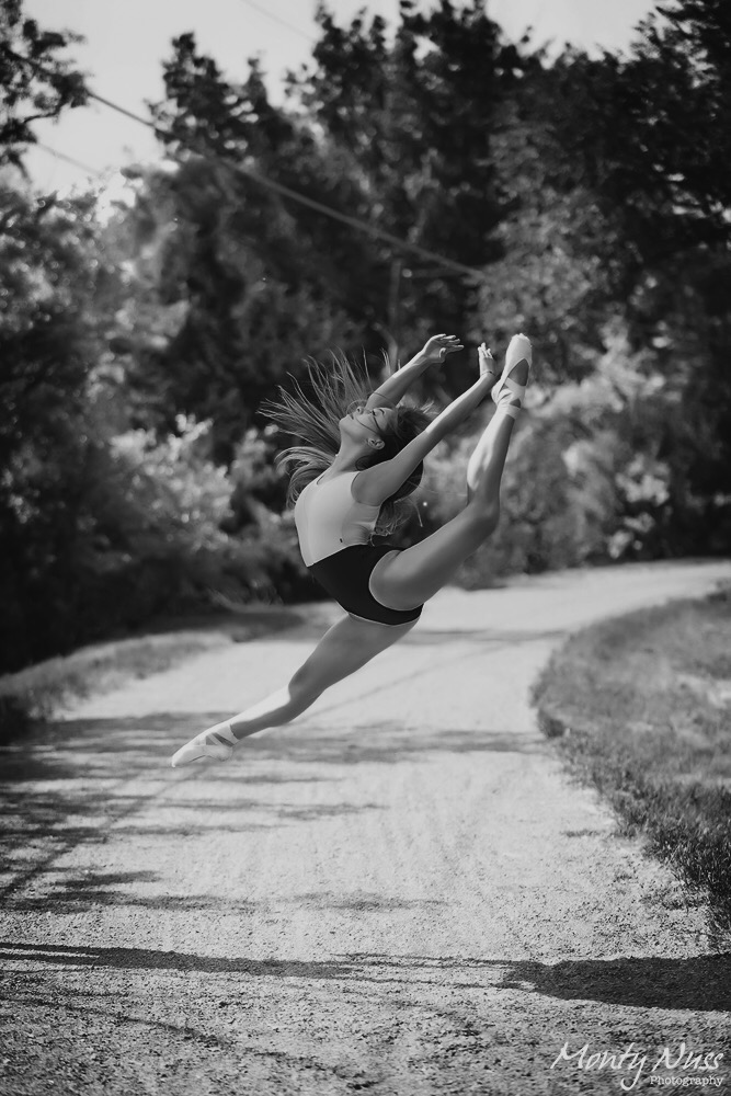 Person mid-air performing a leap on a dirt path surrounded by trees, with hair flowing and arms extended.