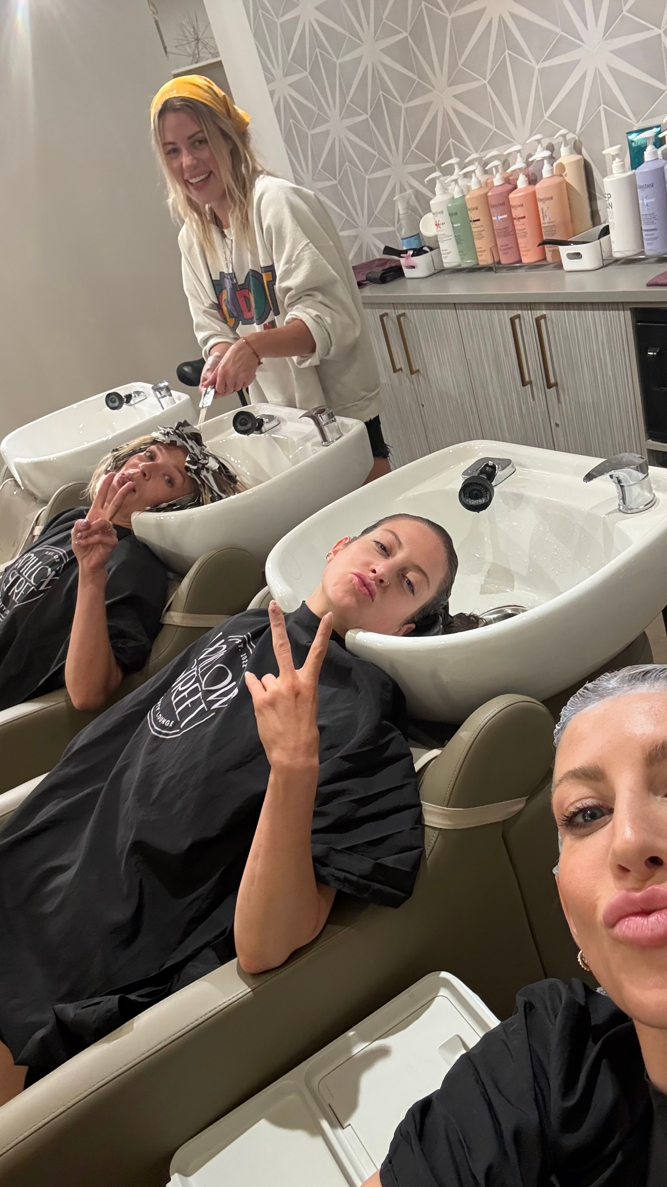 Two women with hair washing stations, one smiling and making a peace sign, the other taking a selfie, in a salon.