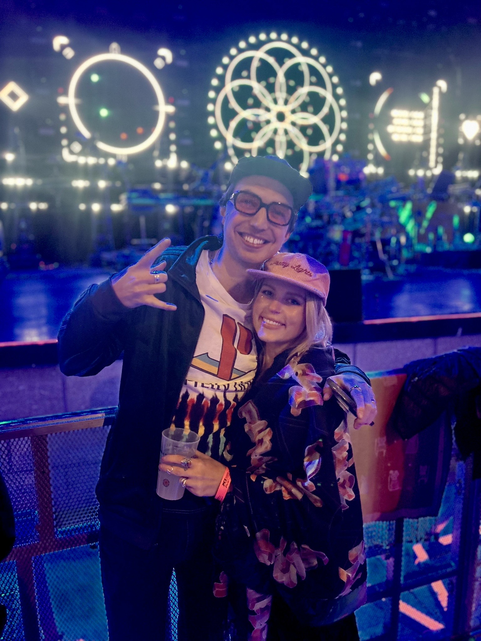 Two people smiling at a nighttime event with bright, colorful lights and decorative patterns in the background.