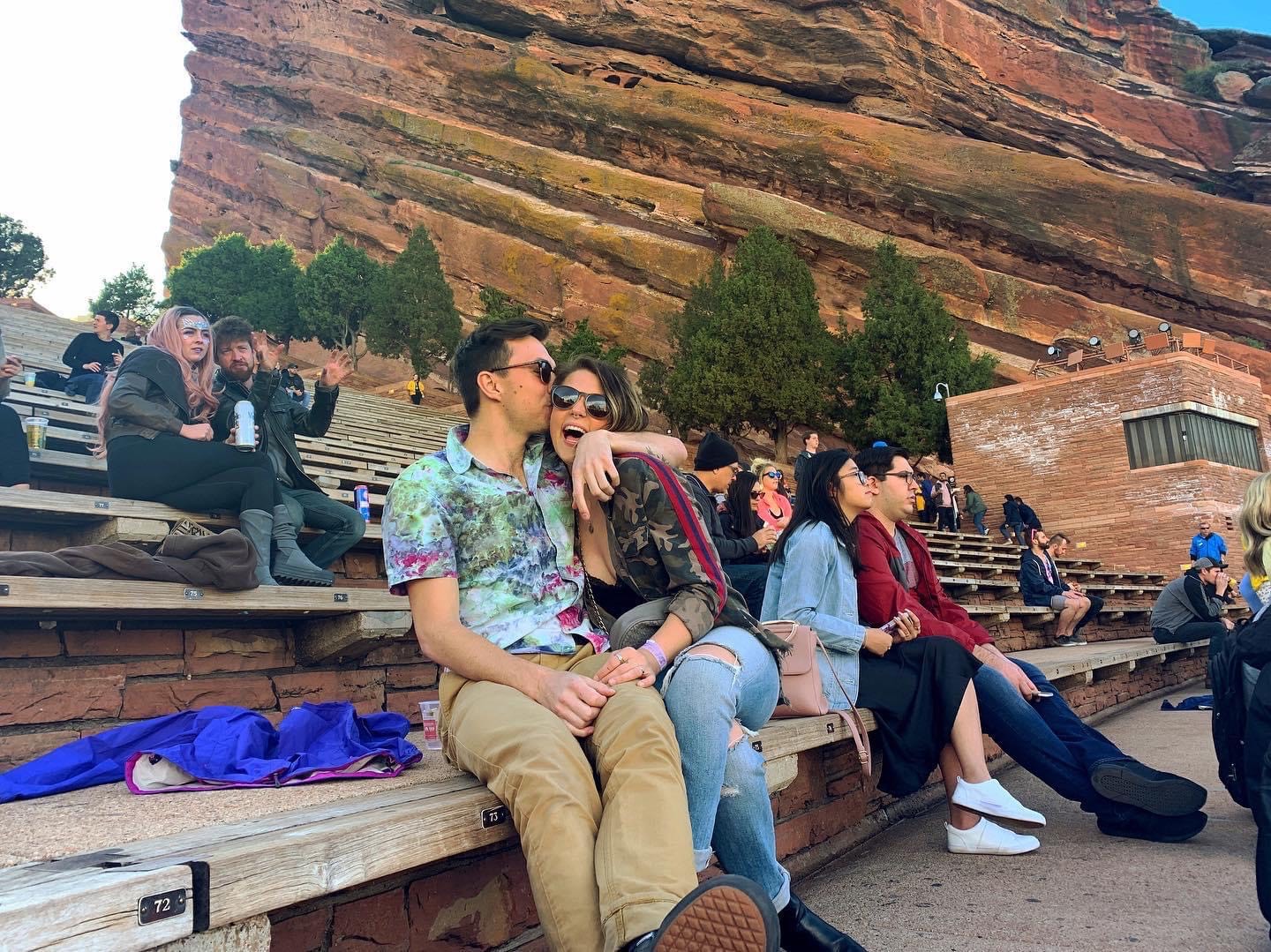 Two people sit close together on a wooden bench, with a rocky hillside and other visitors in the background.