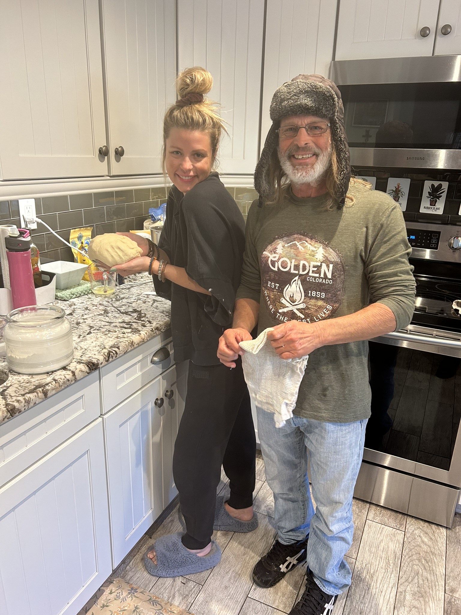 A smiling woman and man stand in a kitchen, preparing food together. The woman is holding a bowl, and the man is holding a cloth.