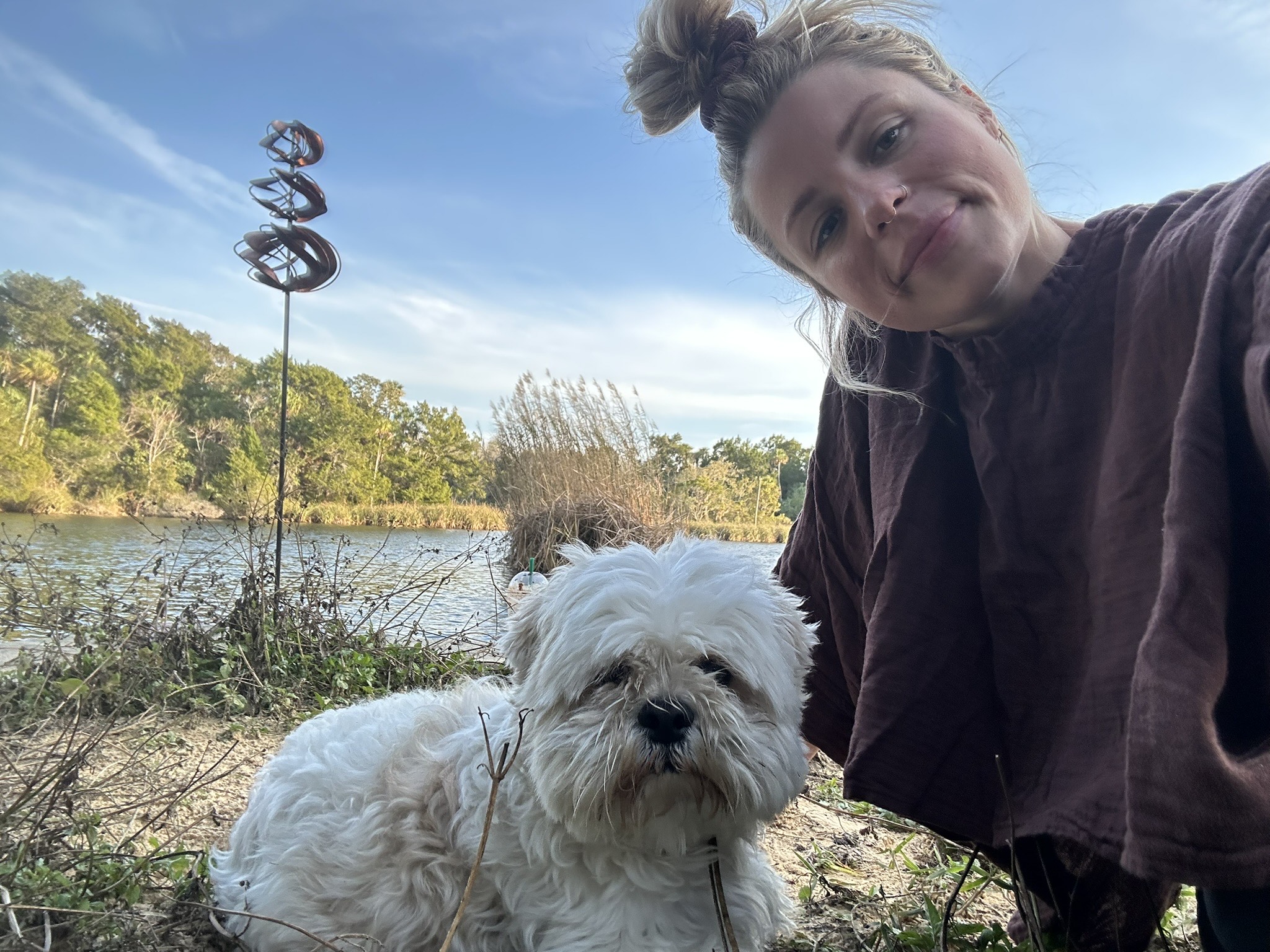 Woman taking a selfie outdoors with a white dog near a body of water and trees in the background.