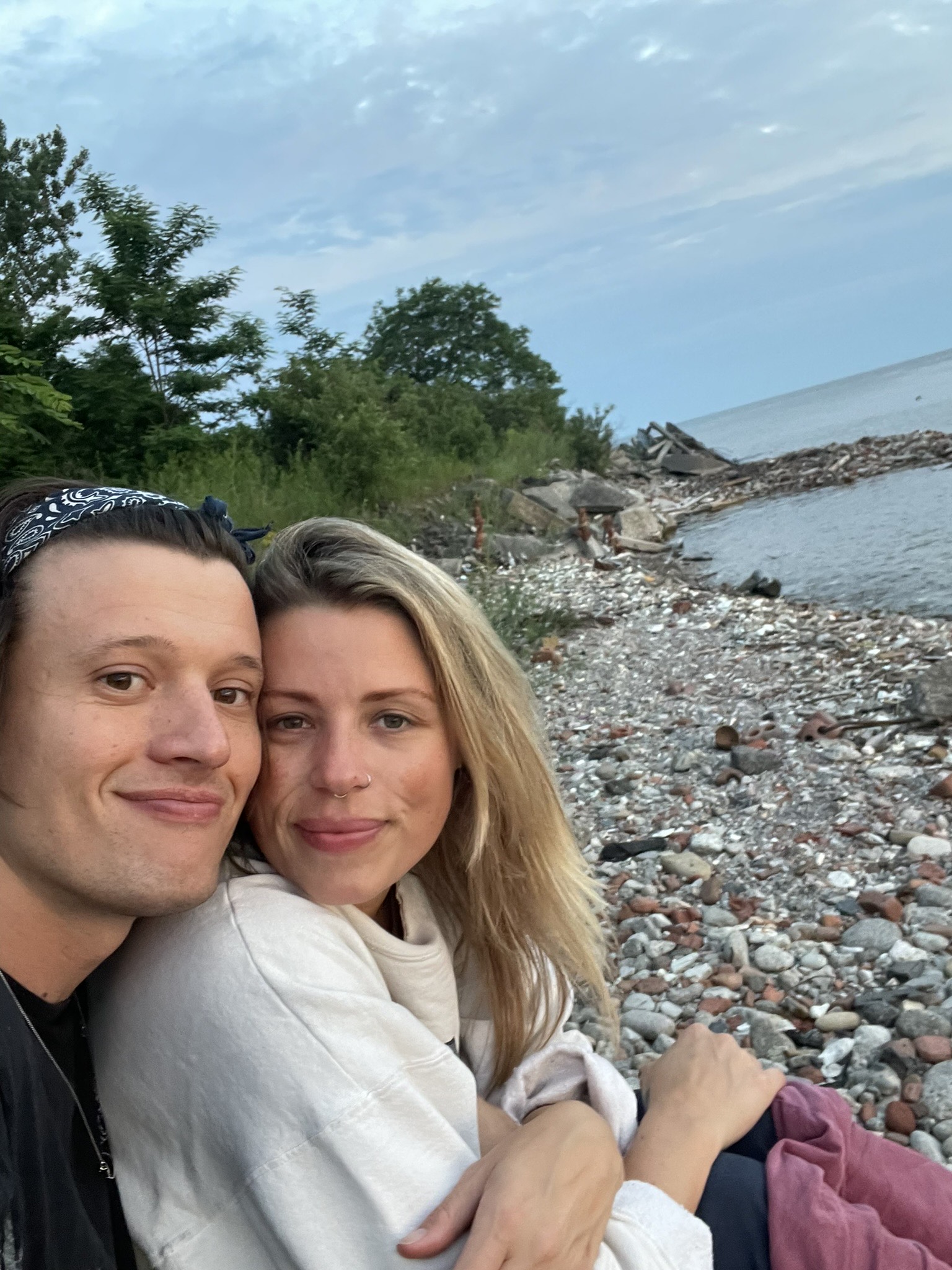 A man and woman sit close together on a rocky shoreline with trees and water in the background.