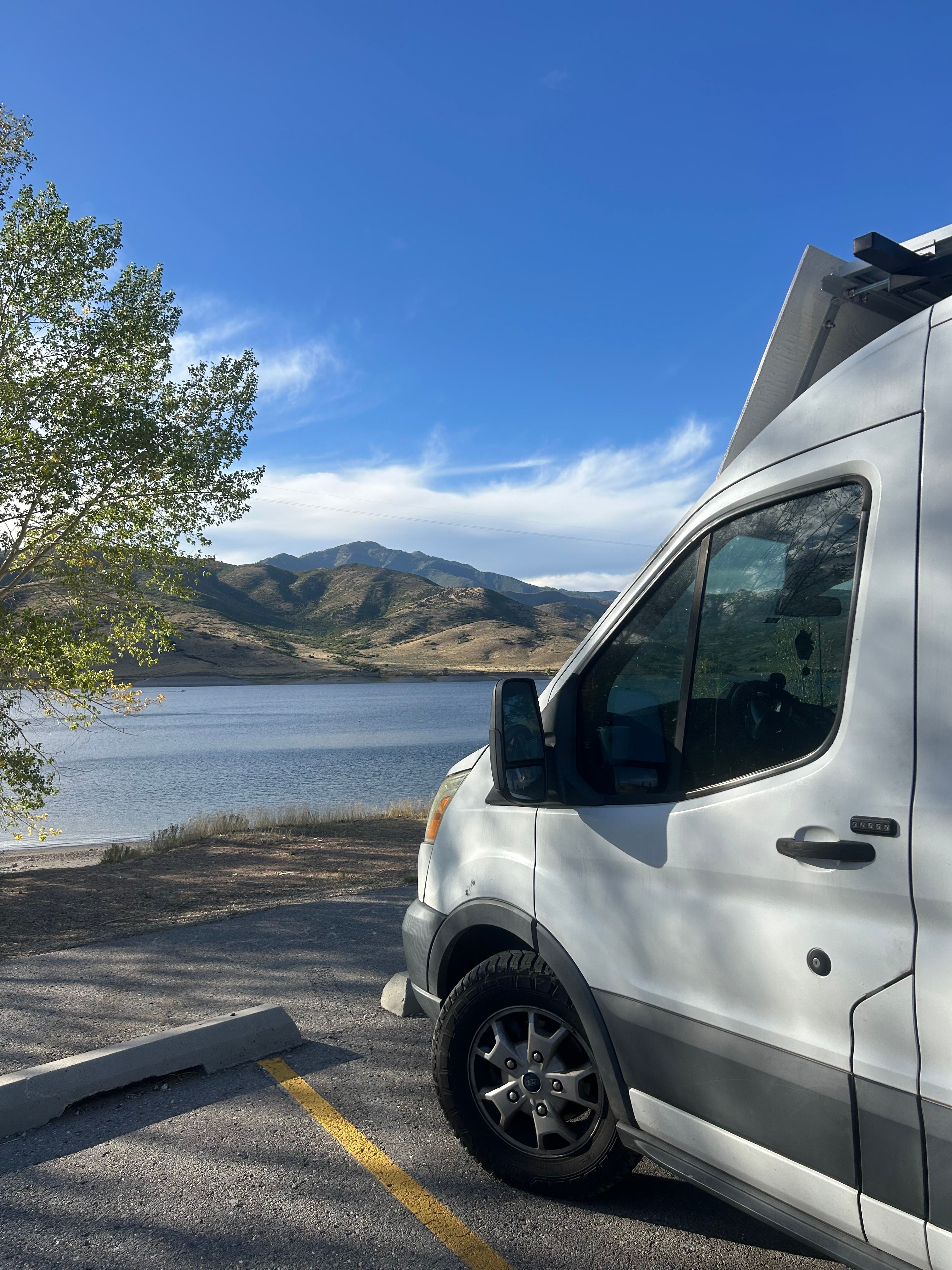 White van parked in a lot near a body of water with mountains in the background and a tree nearby.