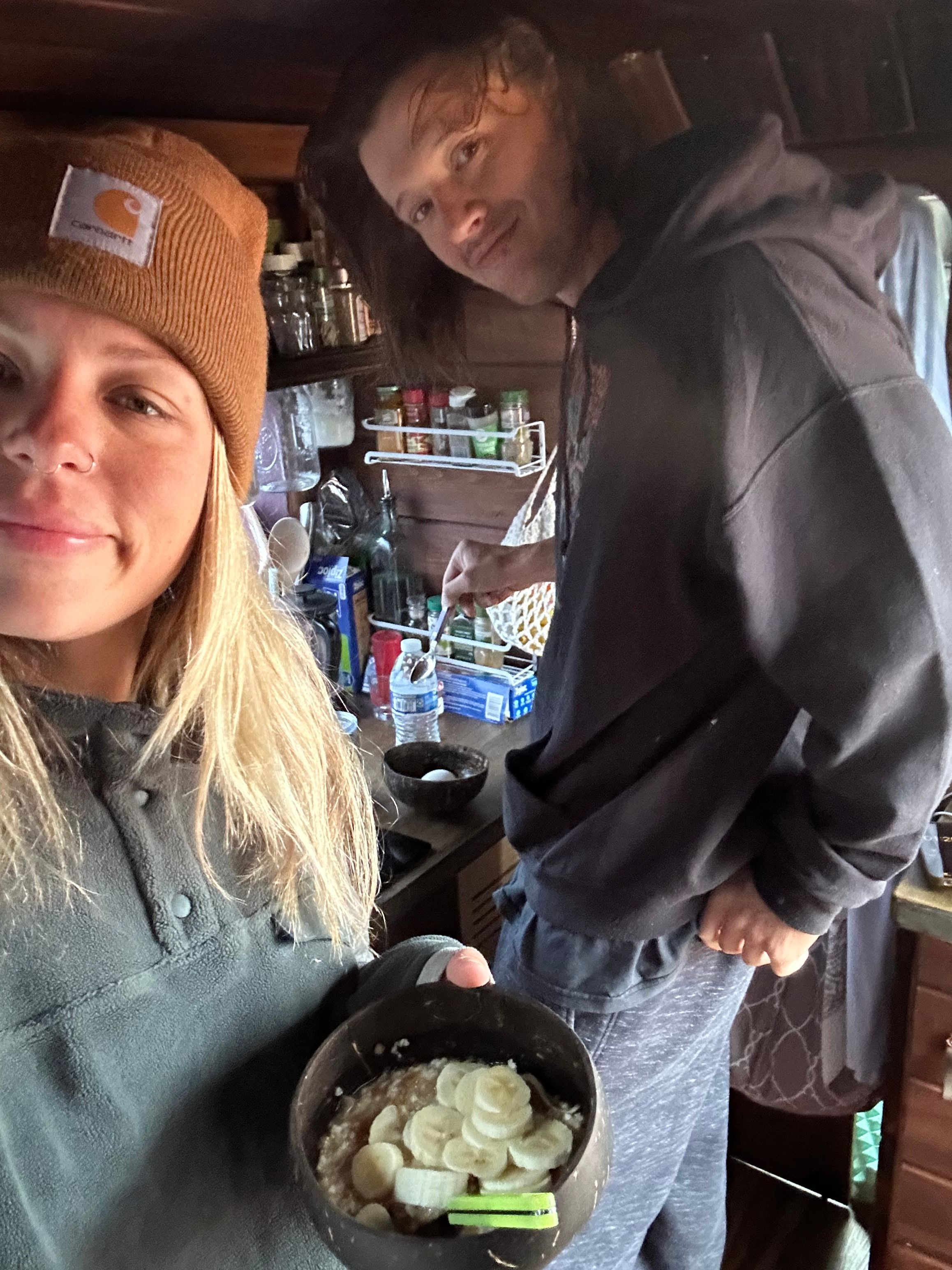 Woman with blonde hair holding a bowl of ice cream, smiling, in a room with a man in the background.