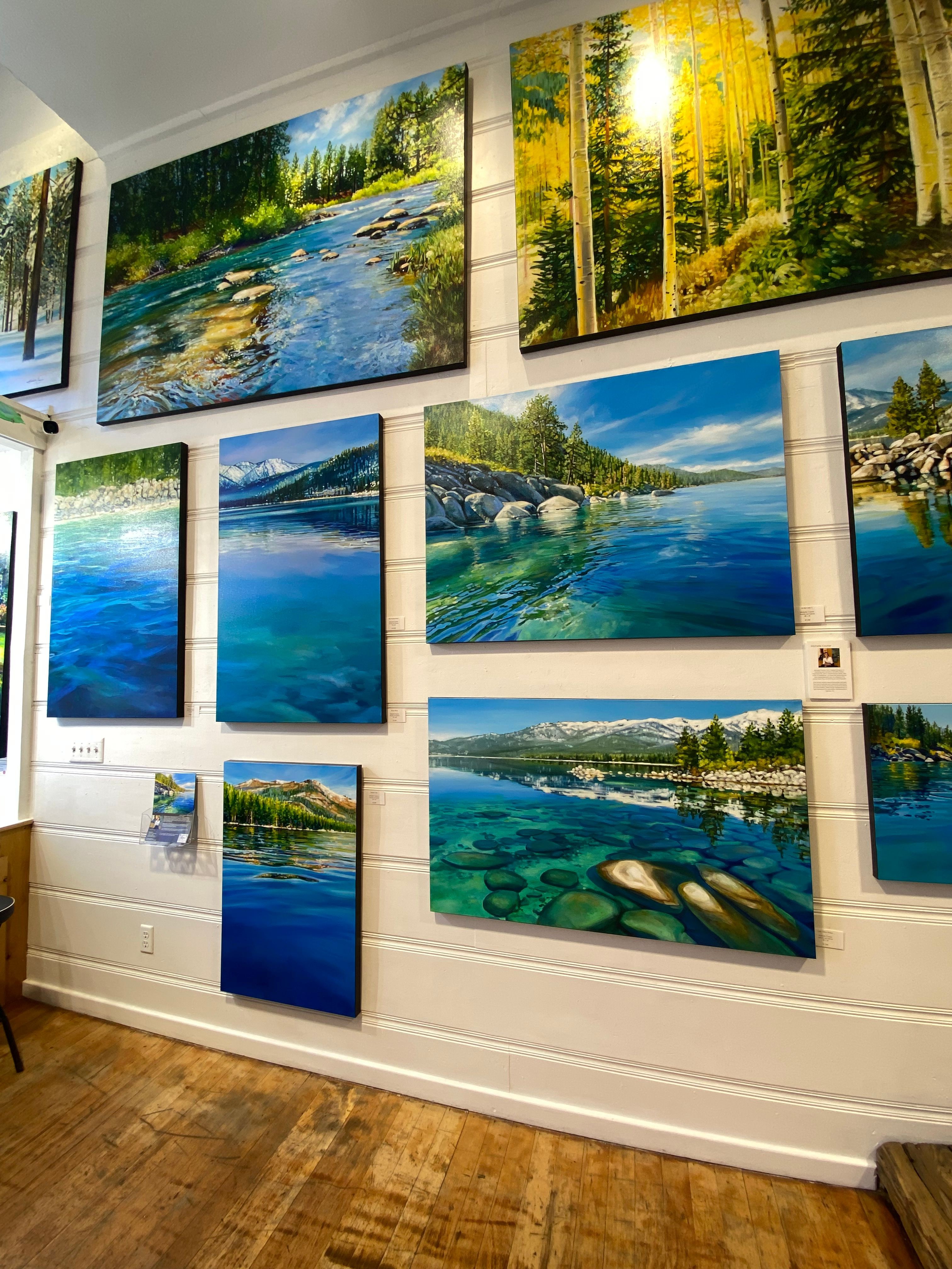 Multiple landscape photographs of lakes, trees, and mountains displayed on a white wall with wooden floor.