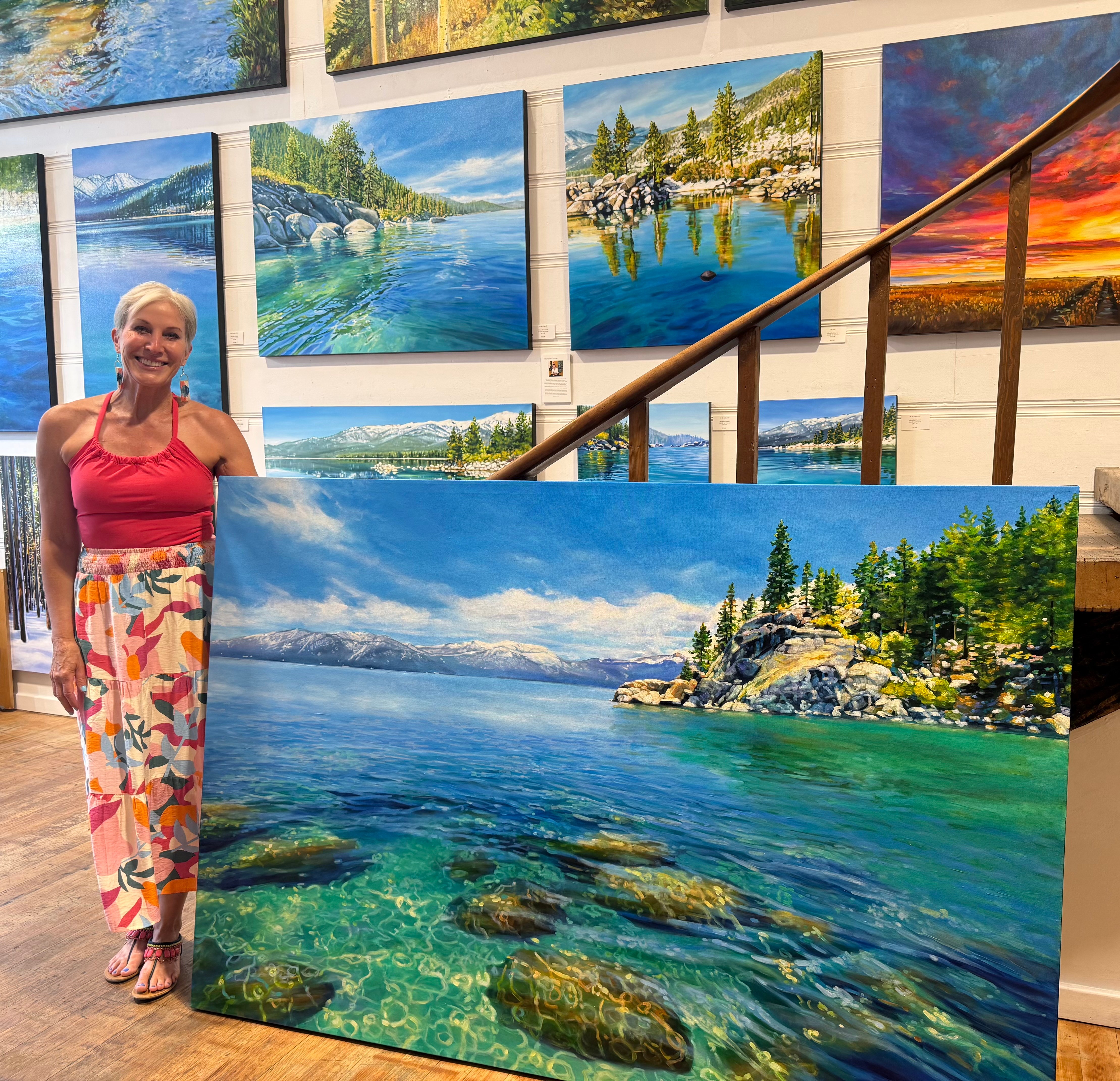 Woman standing next to a large landscape painting of a lake with rocks and trees, in an art gallery with other paintings on the wall.