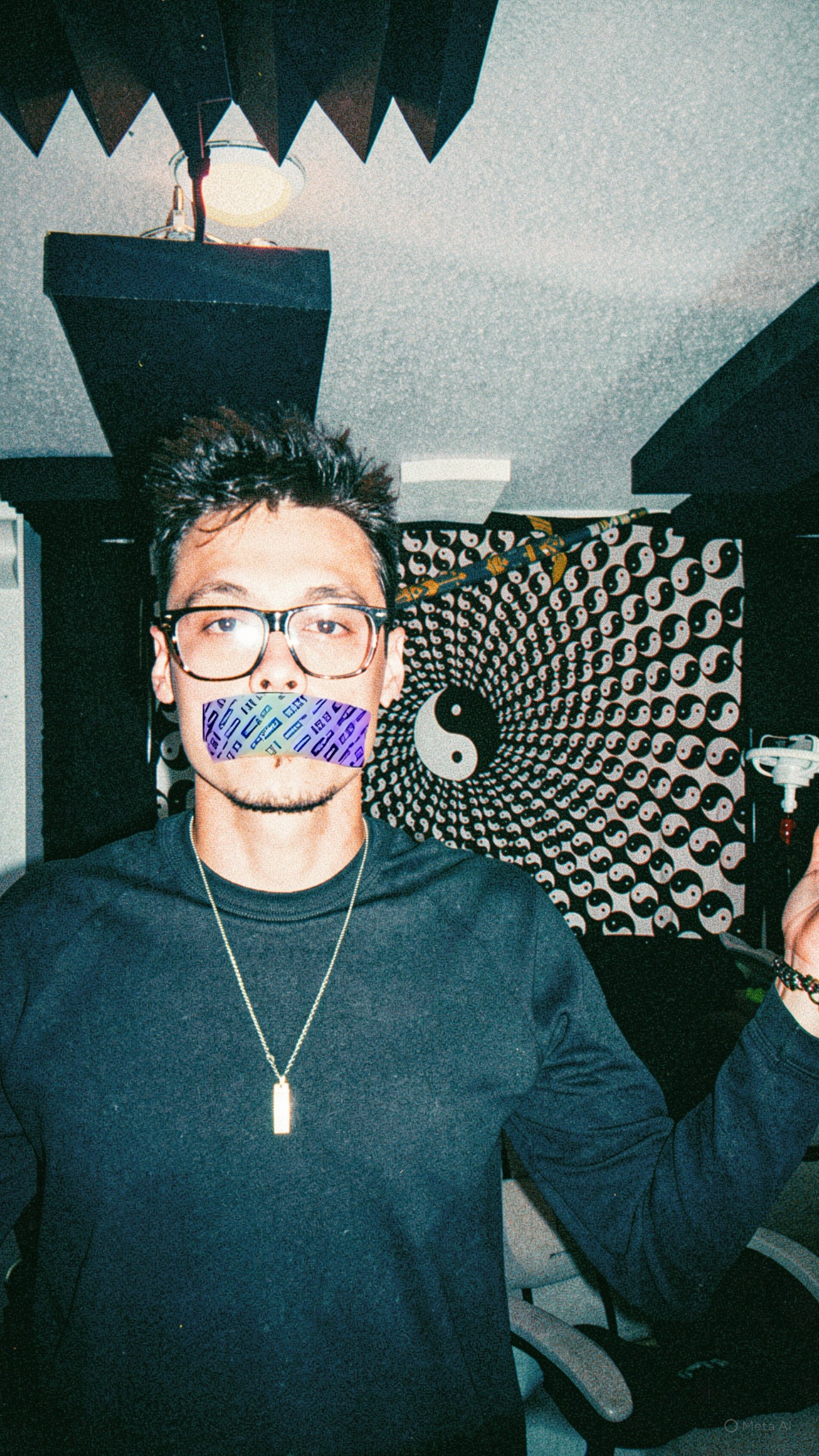 Person with glasses and a necklace, holding an object, standing indoors with patterned wall and ceiling decor behind.