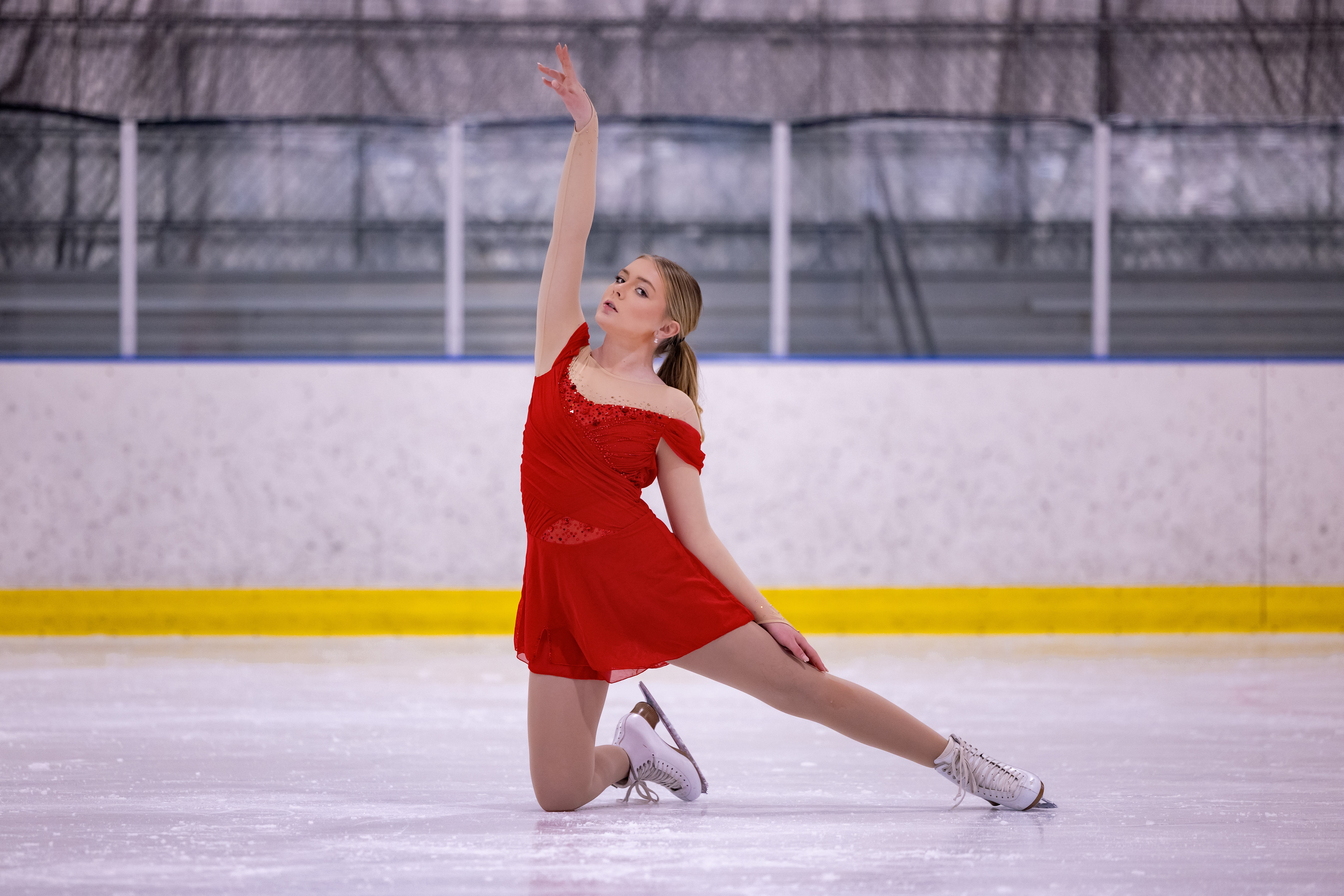 Young girl in a red dress ice skating on an indoor rink, kneeling with one arm raised above her head.