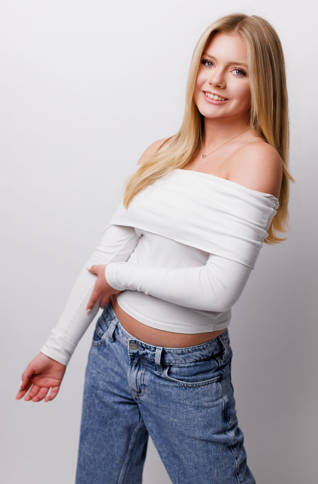 Young woman with long blonde hair smiling, wearing an off-shoulder white top and blue jeans, standing against a plain white background.