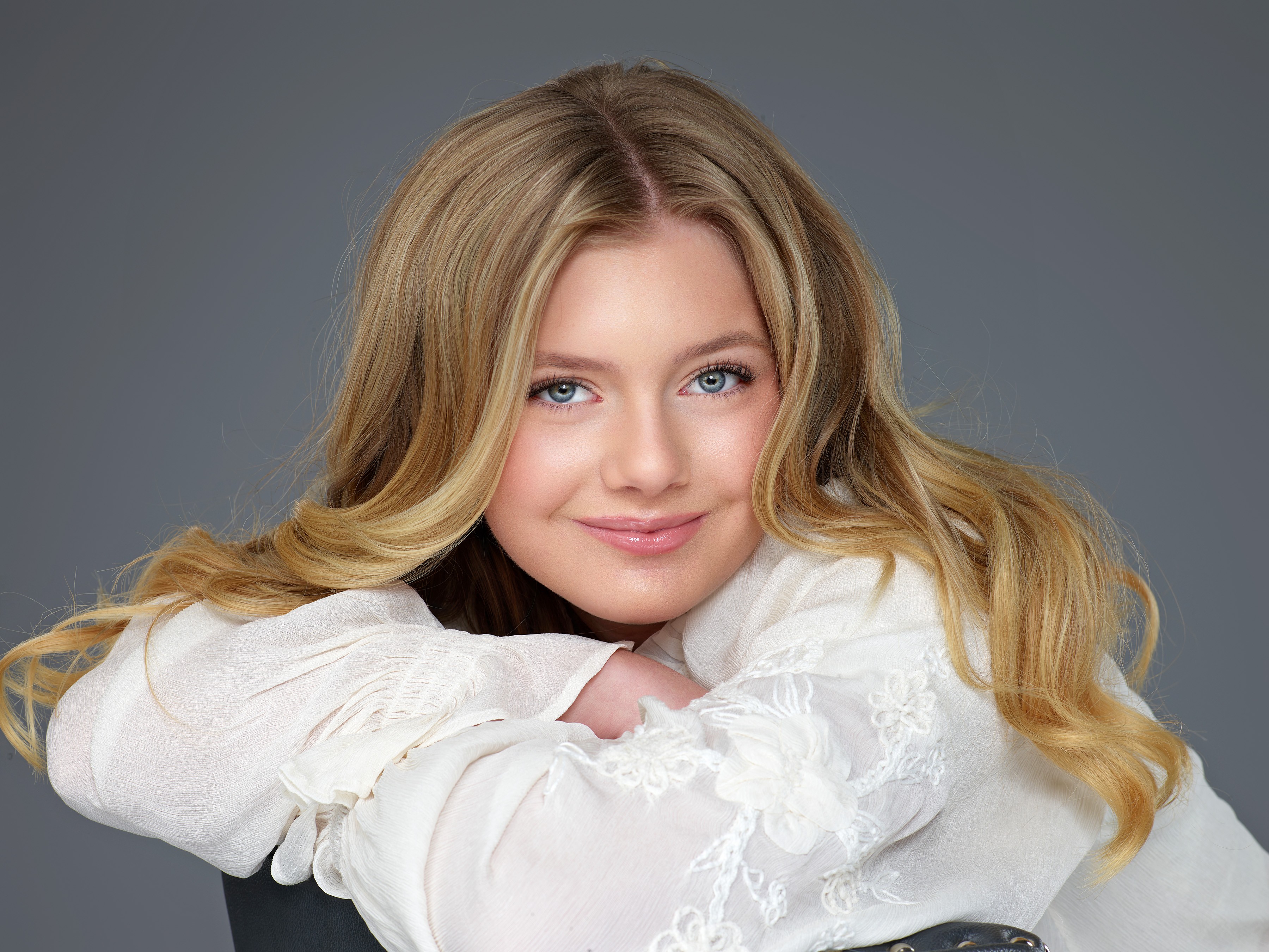 Young woman with long blonde hair and blue eyes, smiling, resting her chin on her crossed arms, wearing a white top, gray background