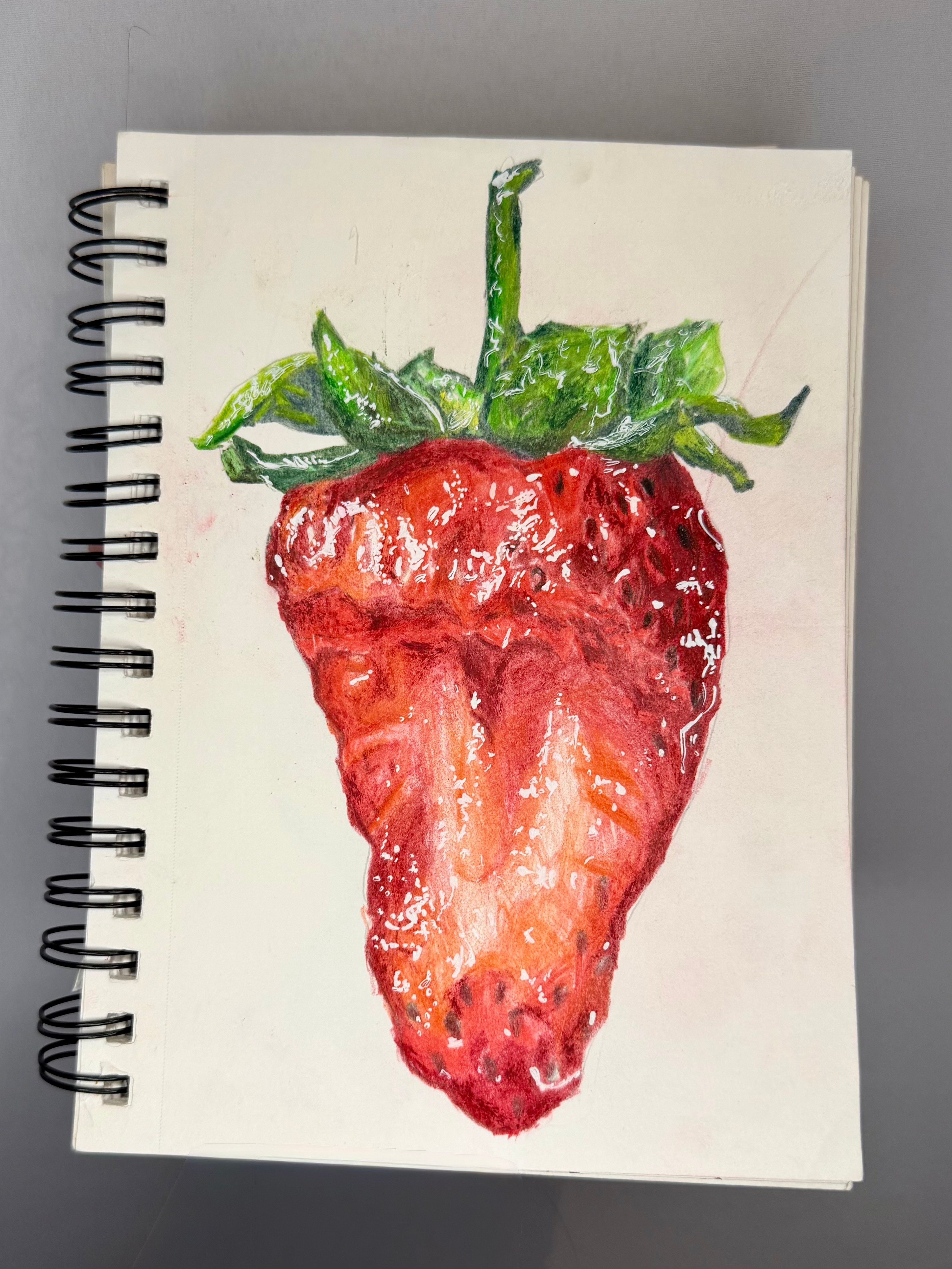 Colorful watercolor painting of a strawberry with green leaves on a white spiral notebook page.