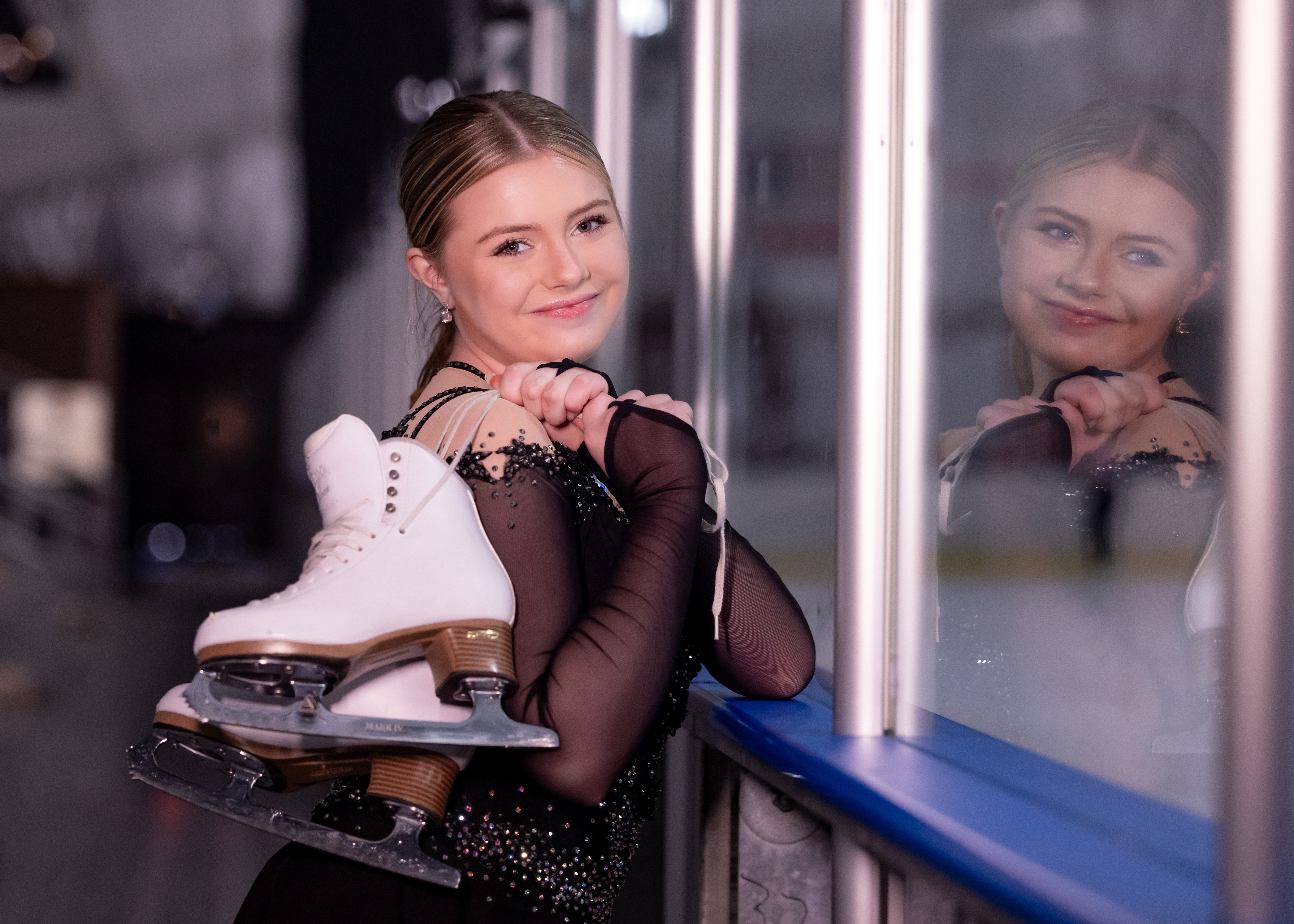 Young woman with ice skates, smiling, leaning on a glass window, reflection visible, indoor setting, evening lighting.