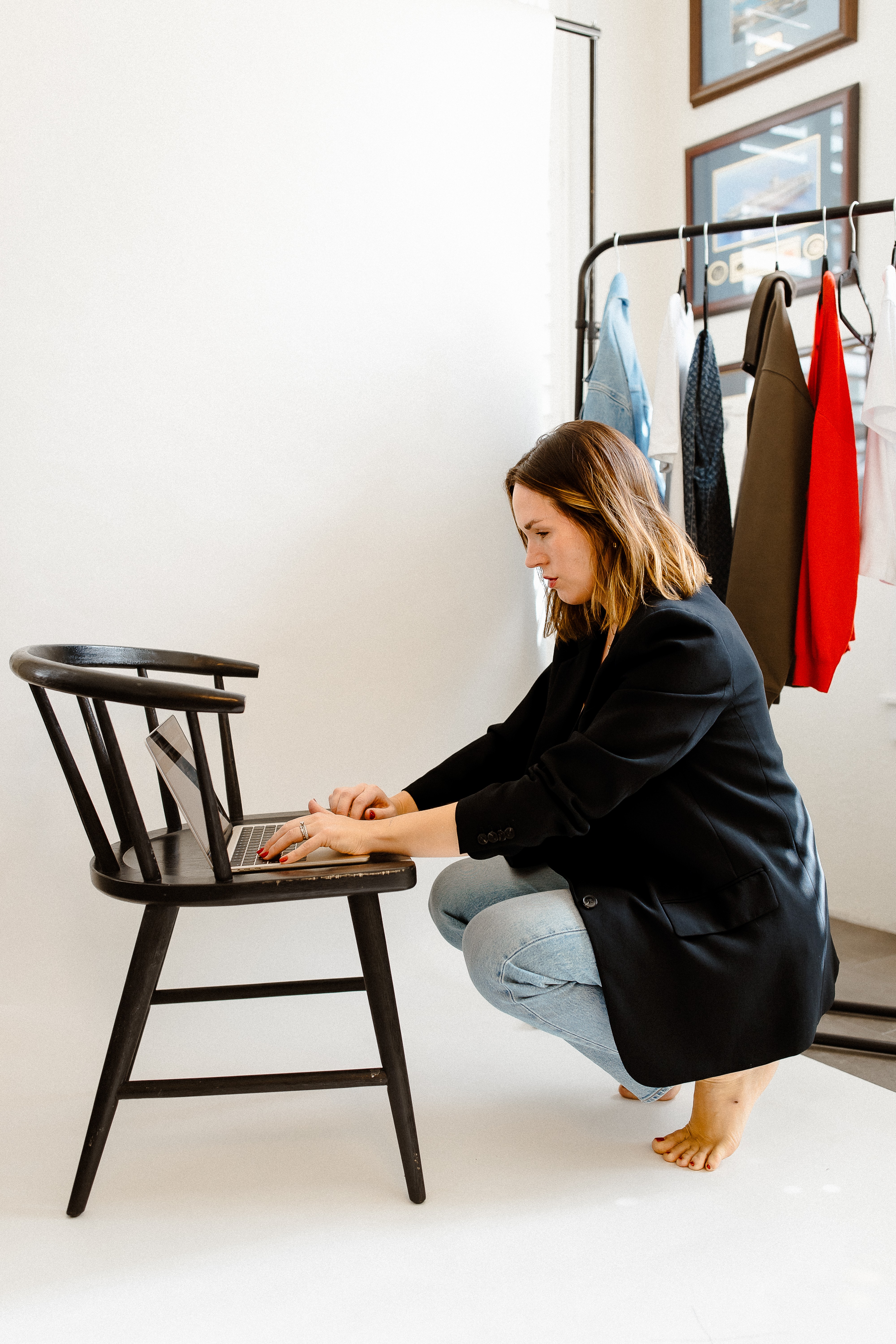 Woman crouching next to a black wooden chair, with clothing hanging on a rack behind her, in a room with framed pictures.