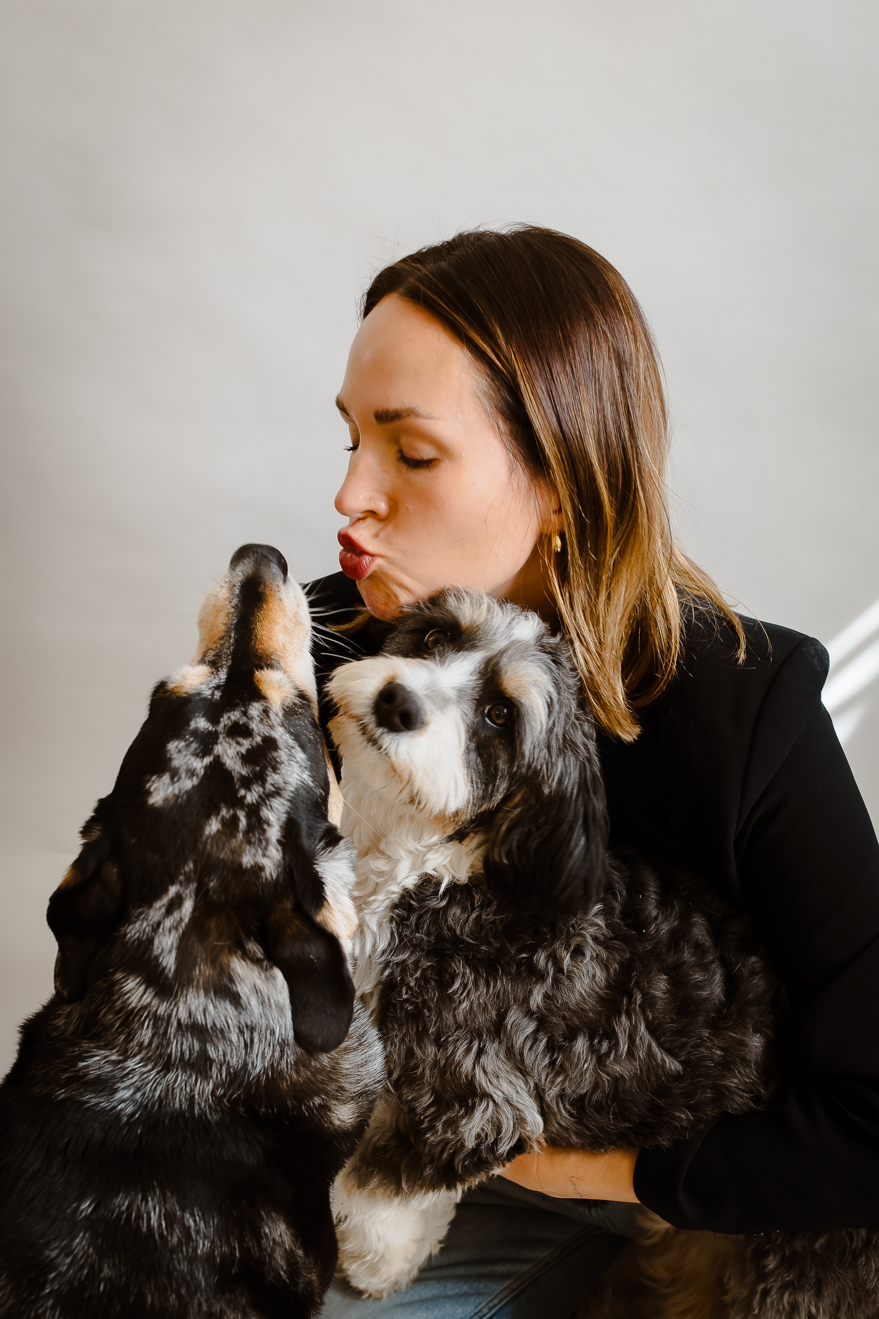 Woman holding two puppies, one licking her face, against a plain background.