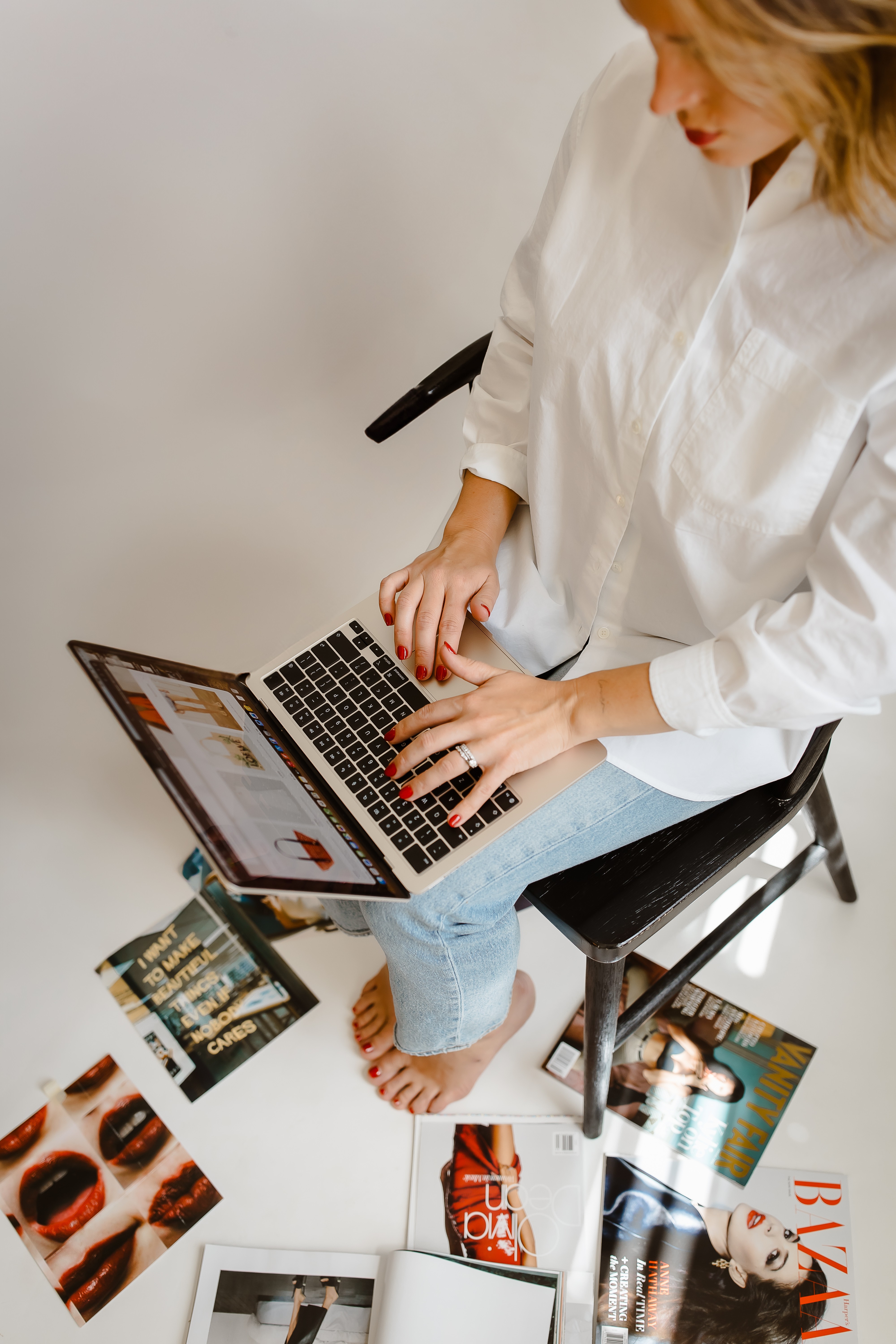 Person sitting on a chair using a laptop, with magazines on the floor around them, in a white room.