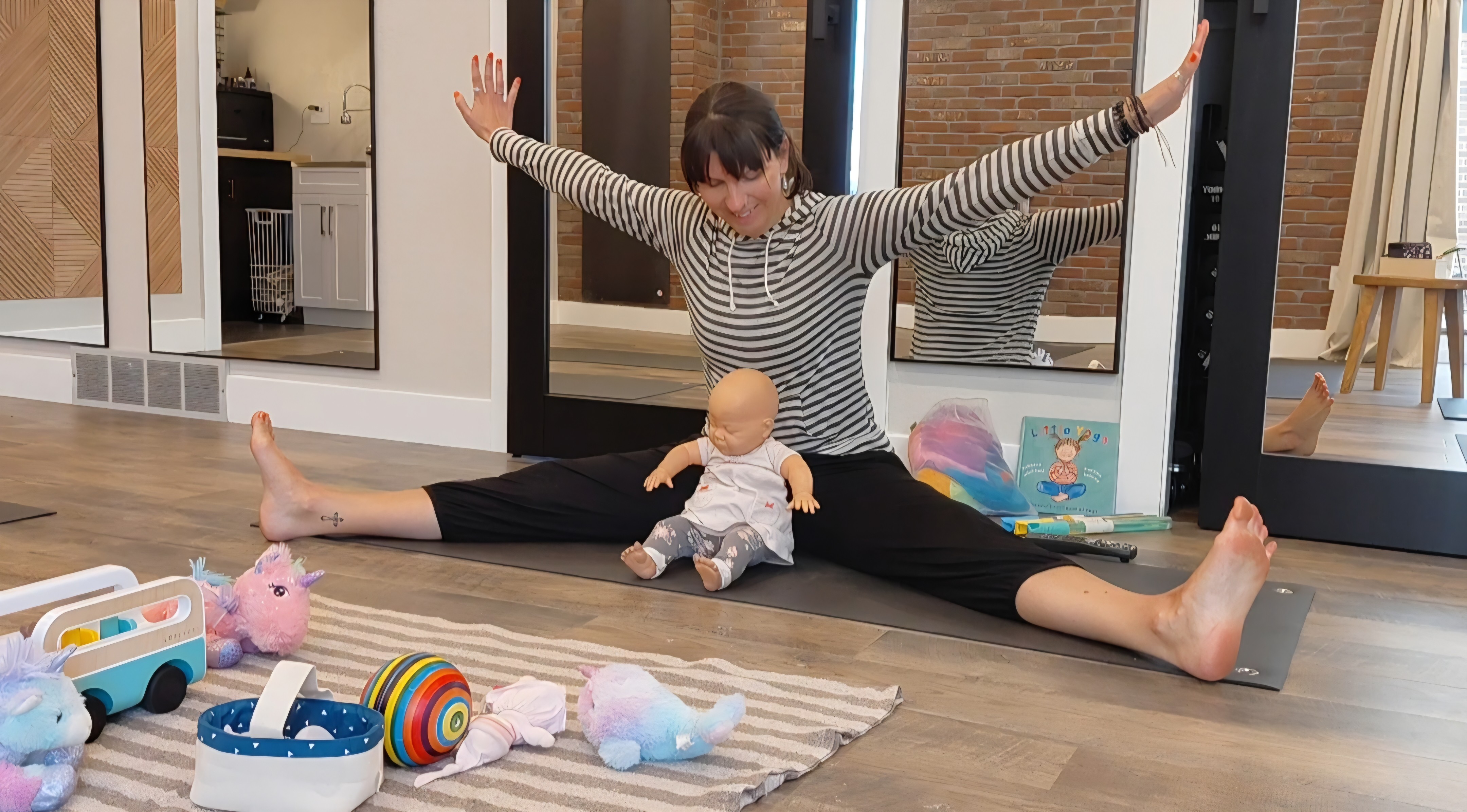 Person sitting on the floor with legs spread wide, holding a baby doll, surrounded by toys, indoors.