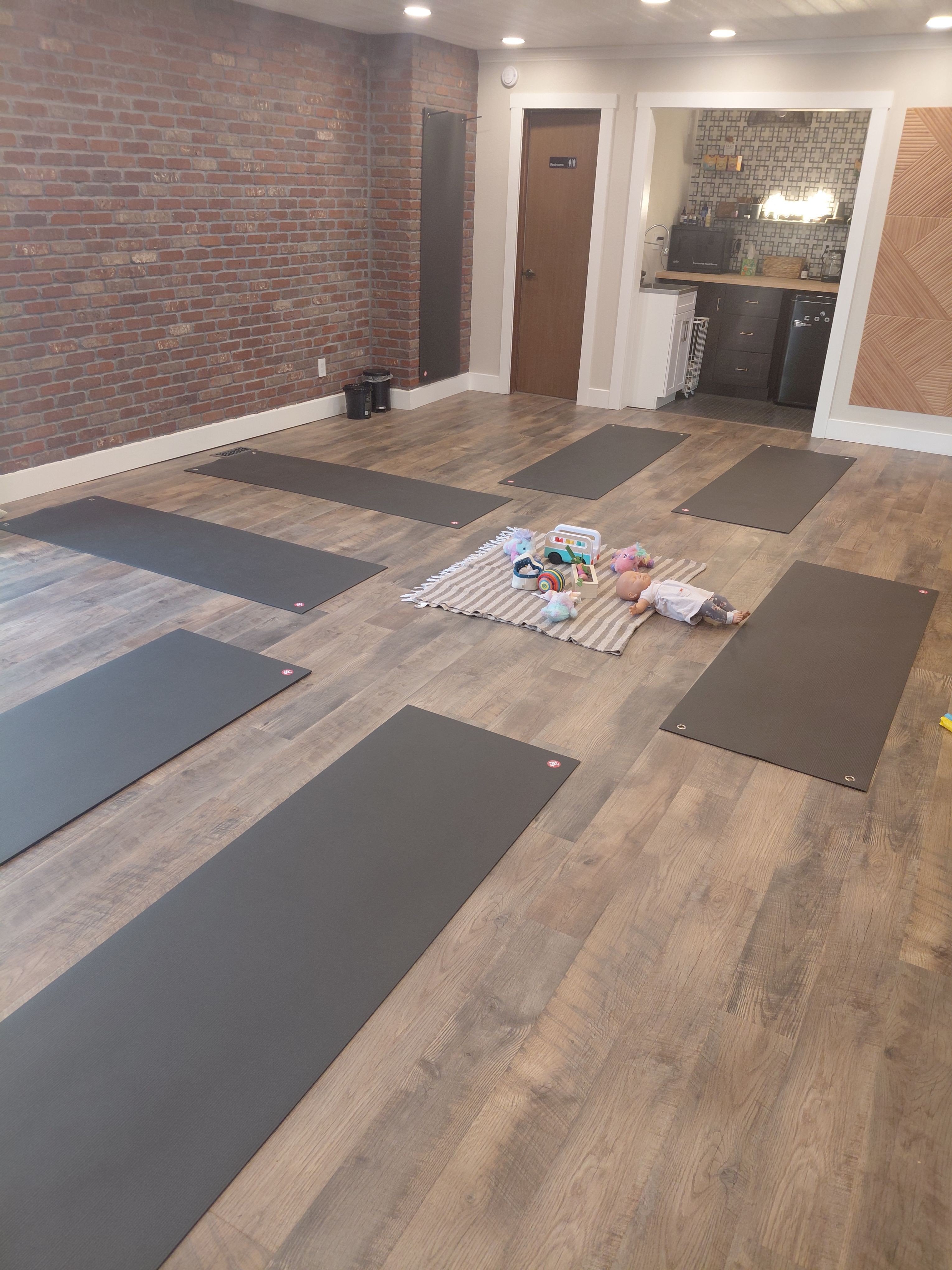 Room with wooden floor, brick wall, and yoga mats arranged on the floor, with a door and window in the background.