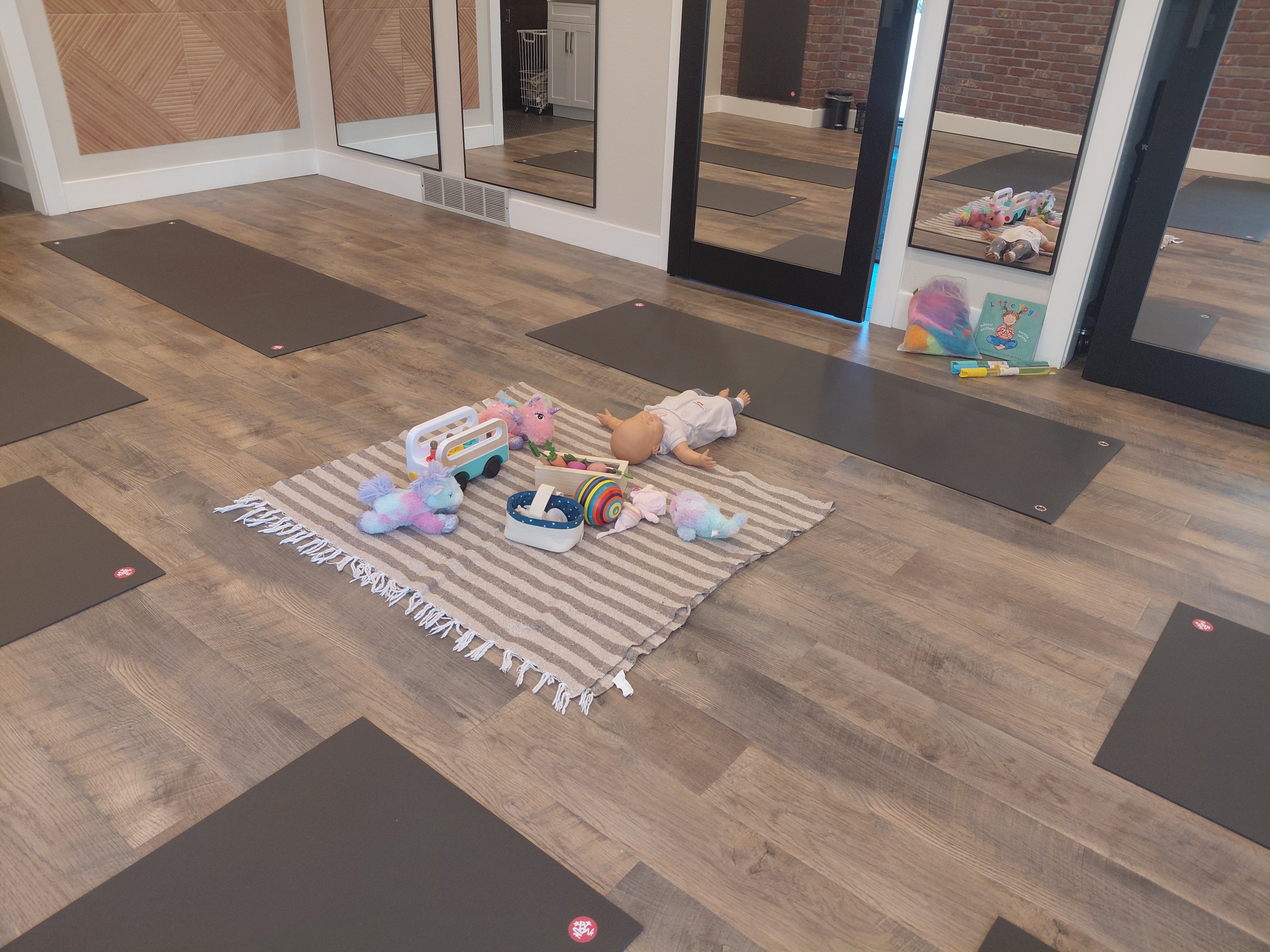 Room with wooden floor, yoga mats, and a striped rug with toys and stuffed animals, glass doors in background.